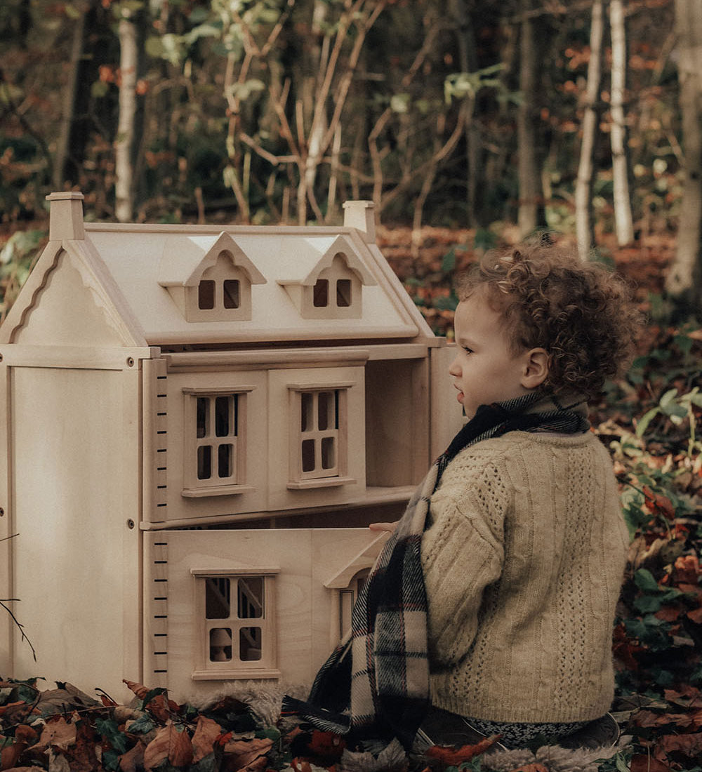 A child sitting on a leaf covered floor in the forest playing with a PlanToys Victorian Dolls House.
