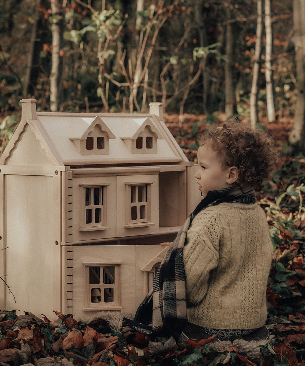 A child sitting on a leaf covered floor in the forest playing with a PlanToys Victorian Dolls House.
