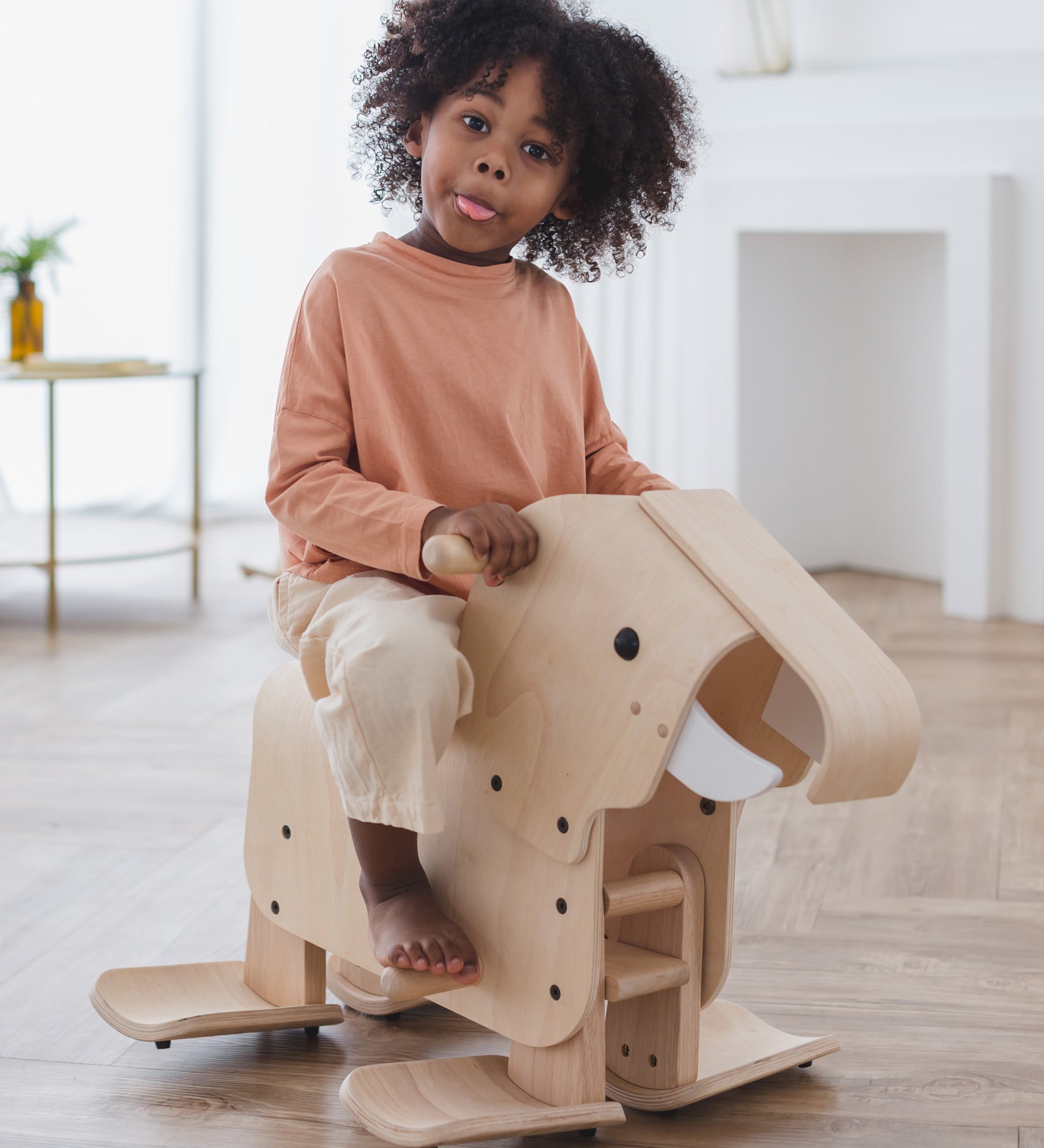 A child riding on a PlanToys Walking Elephant, they have a playful expression and are sticking their tongue out. 