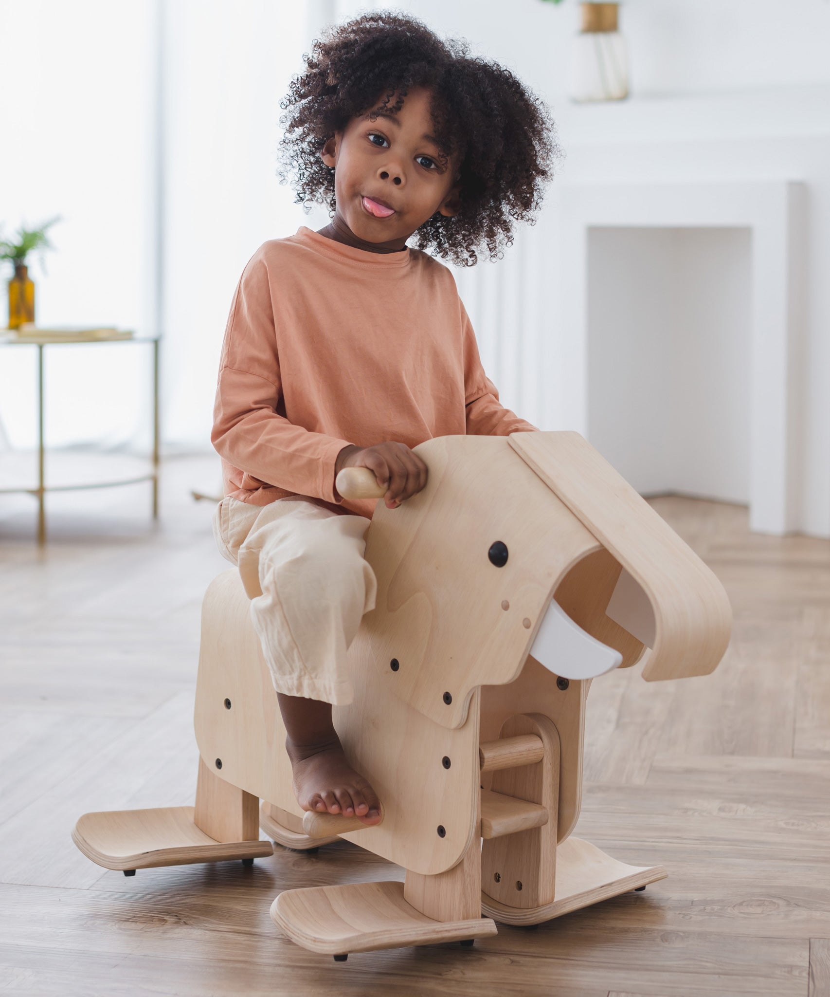 A child riding on a PlanToys Walking Elephant, they have a playful expression and are sticking their tongue out. 