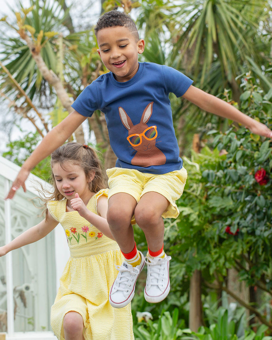  wearing the Frugi Carsen Applique T-Shirt - Hare. A deep navy blue GOTS organic cotton t-shirt with a brown hare wearing yellow sunglasses with a rainbow reflection in the lens. The child is also wearing Yell