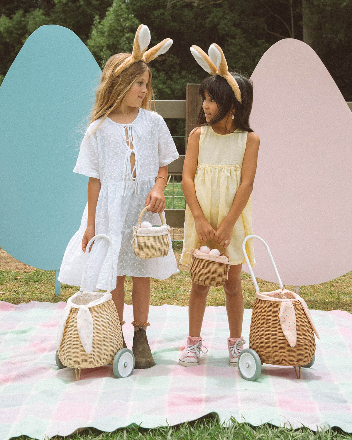 A child smiling and holding an Olli Ella Lamby Doll. In front of the child is an Olli Ella Rattan Lined Basket in Gumdrop