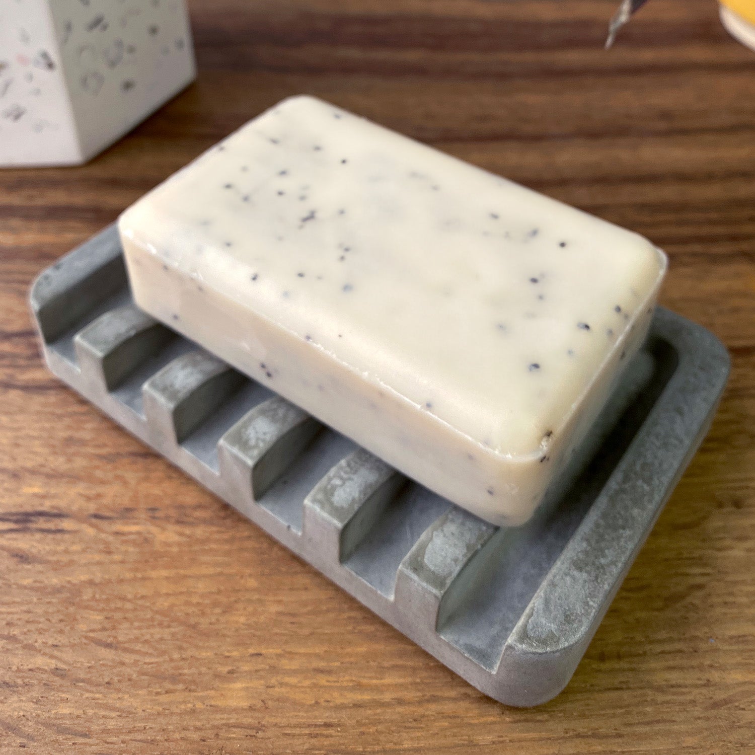 Rawr Grey Concrete Soap Dish on a wooden shelf. There is a soap bar resting on the dish. In the background is a Rawr Shel Terrazzo Storage pot.