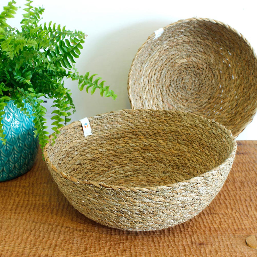 Large and medium respiin woven seagrass bowls on a tan placeman in front of a while wall next to a plant