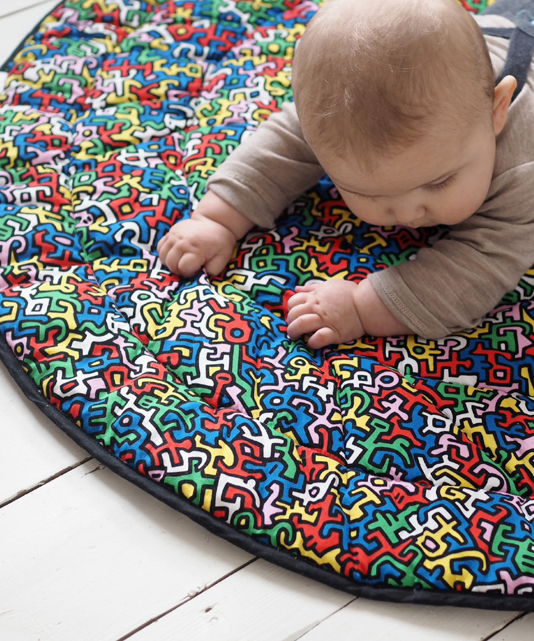 A baby lying down and looking at the 'Baby' print on the Etta Loves X Keith Haring Reversible Organic Cotton Baby Play Mat