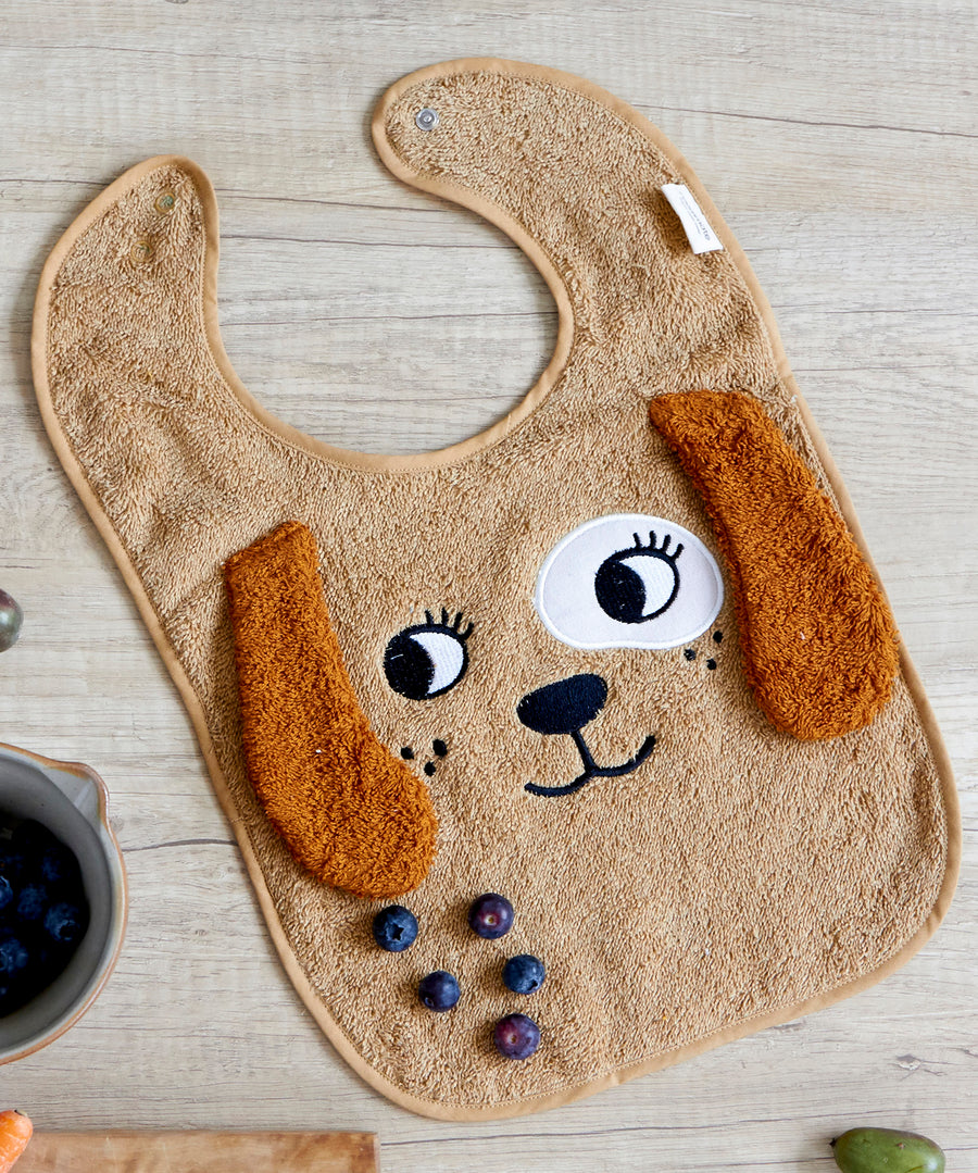 The Roommate Dog Baby Bib with an embroidered puppy face with floppy 3D ears. On a tabletop with some blueberries. 