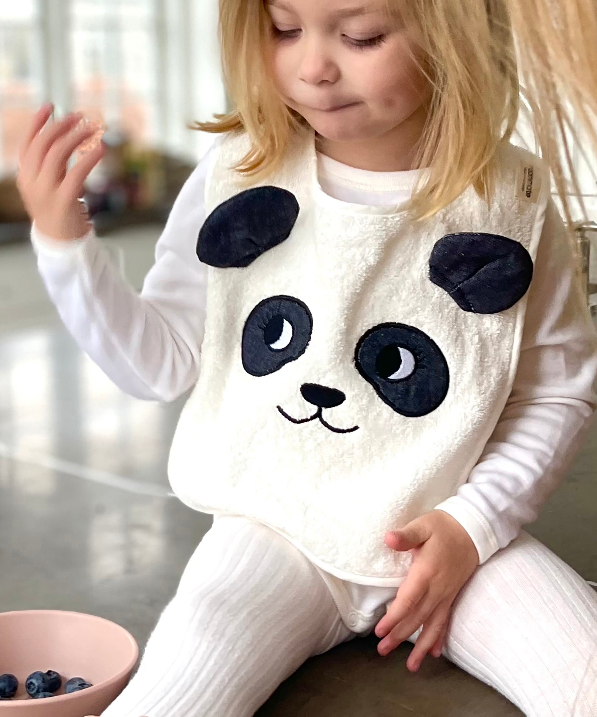 Child sat on a grey kitchen worktop wearing the Roommate Panda Baby Bib in organic cotton with embroidered panda face with 3D ears. She has a bowl of blueberries beside her.