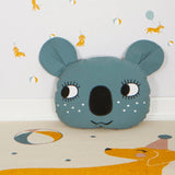 Close up of a Roommate organic cotton blue koala bear cushion on a beige carpet in front of a dog patterned wallpaper