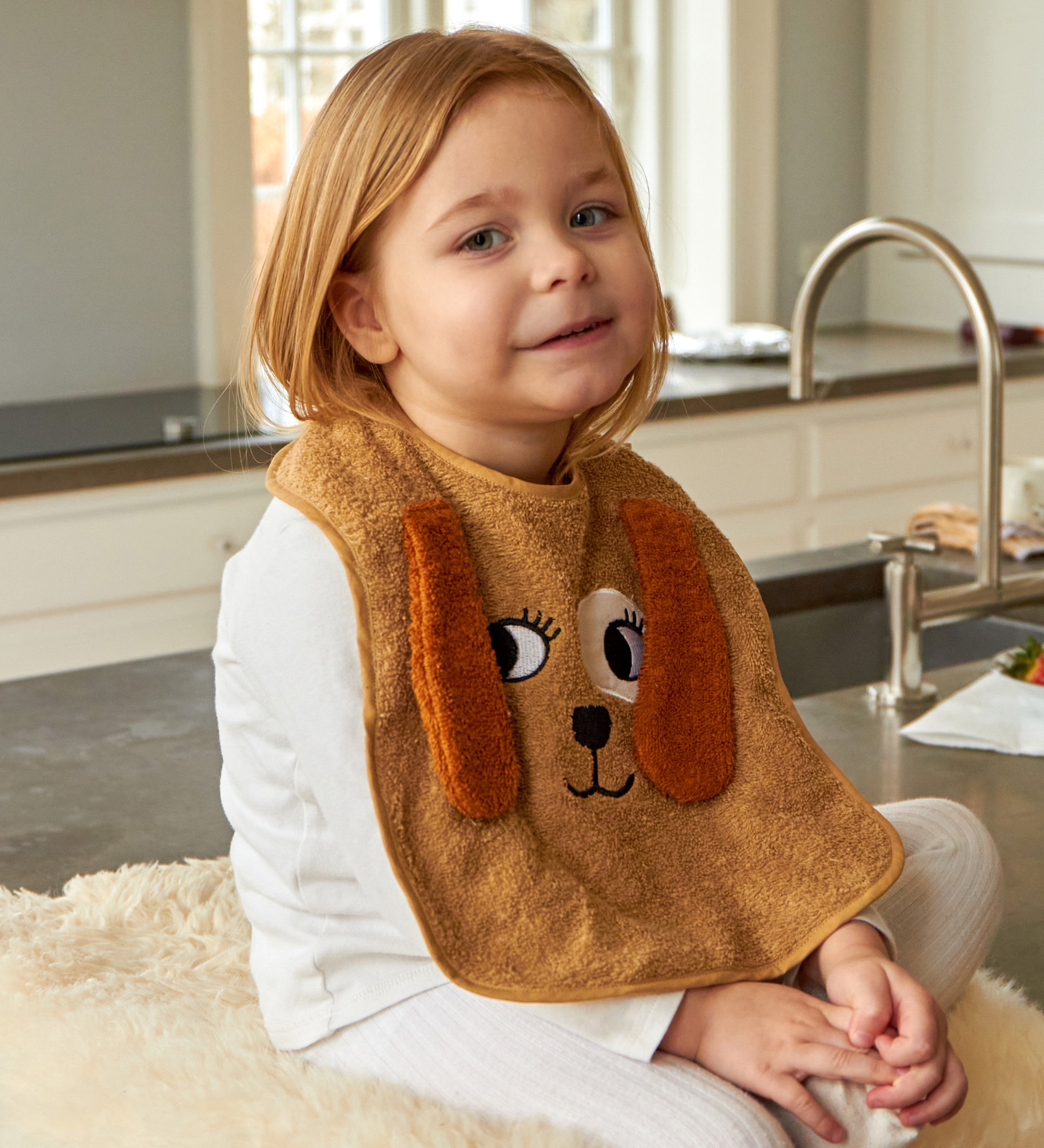 Child wearing the Roommate Dog Baby Bib with an embroidered puppy face with floppy 3D ears. She is sat on a worktop on a fleece rug.