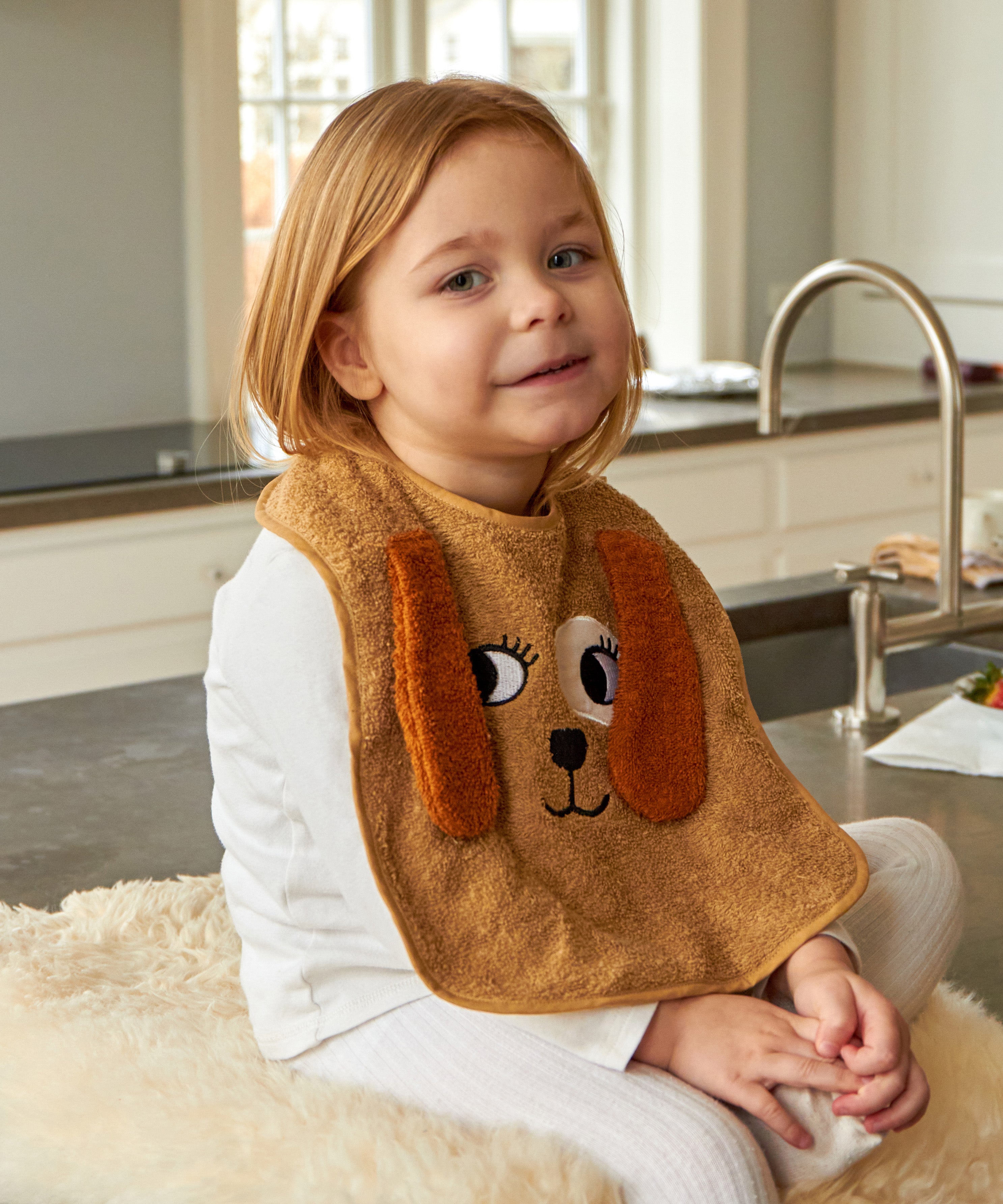 Child wearing the Roommate Dog Baby Bib with an embroidered puppy face with floppy 3D ears. She is sat on a worktop on a fleece rug.