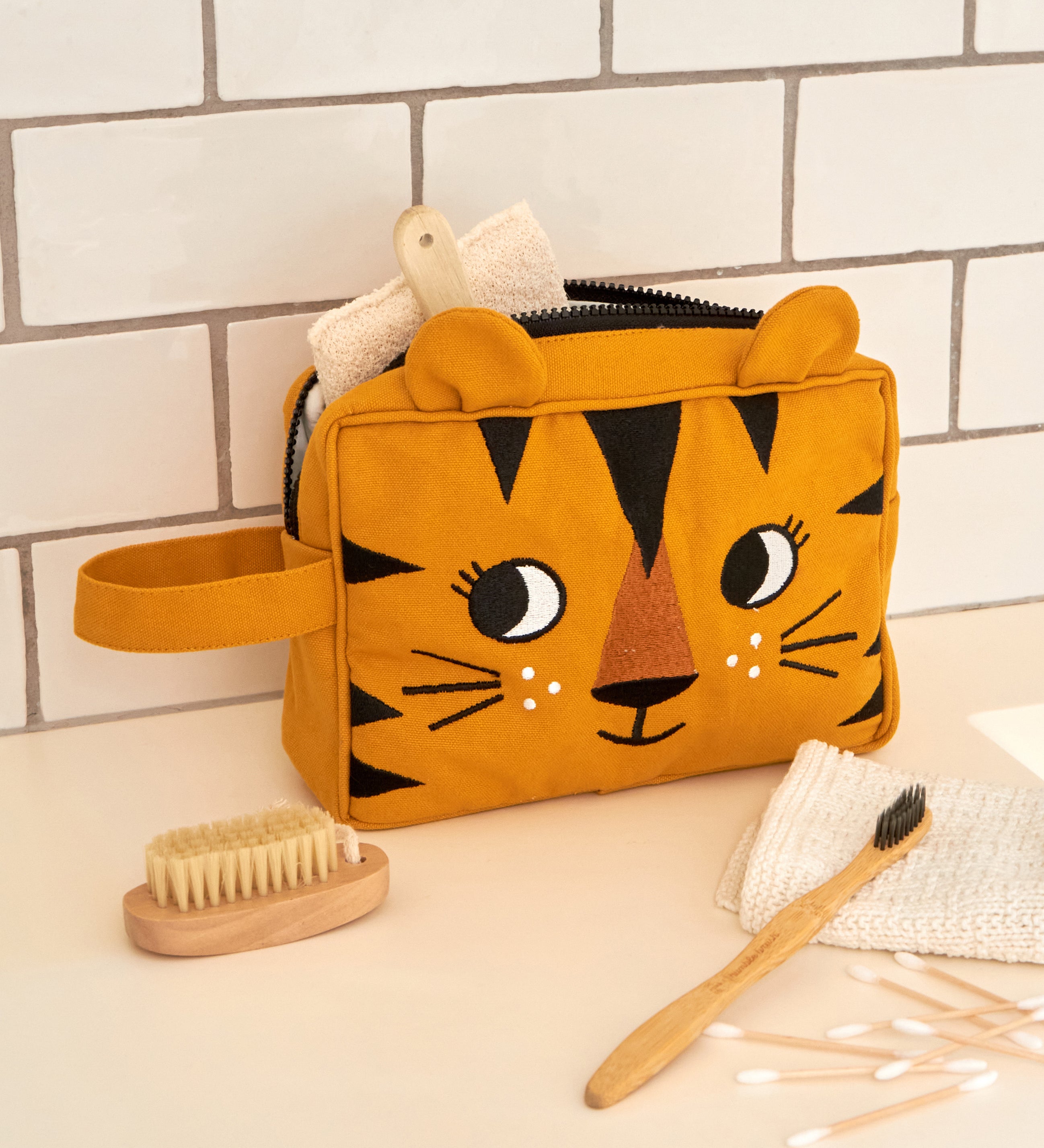 The Roommate organic cotton kids washbag, in a sunny yellow with an embroidered tiger design and 3D tiger ears. It is in a bathroom and filled with toiletries.