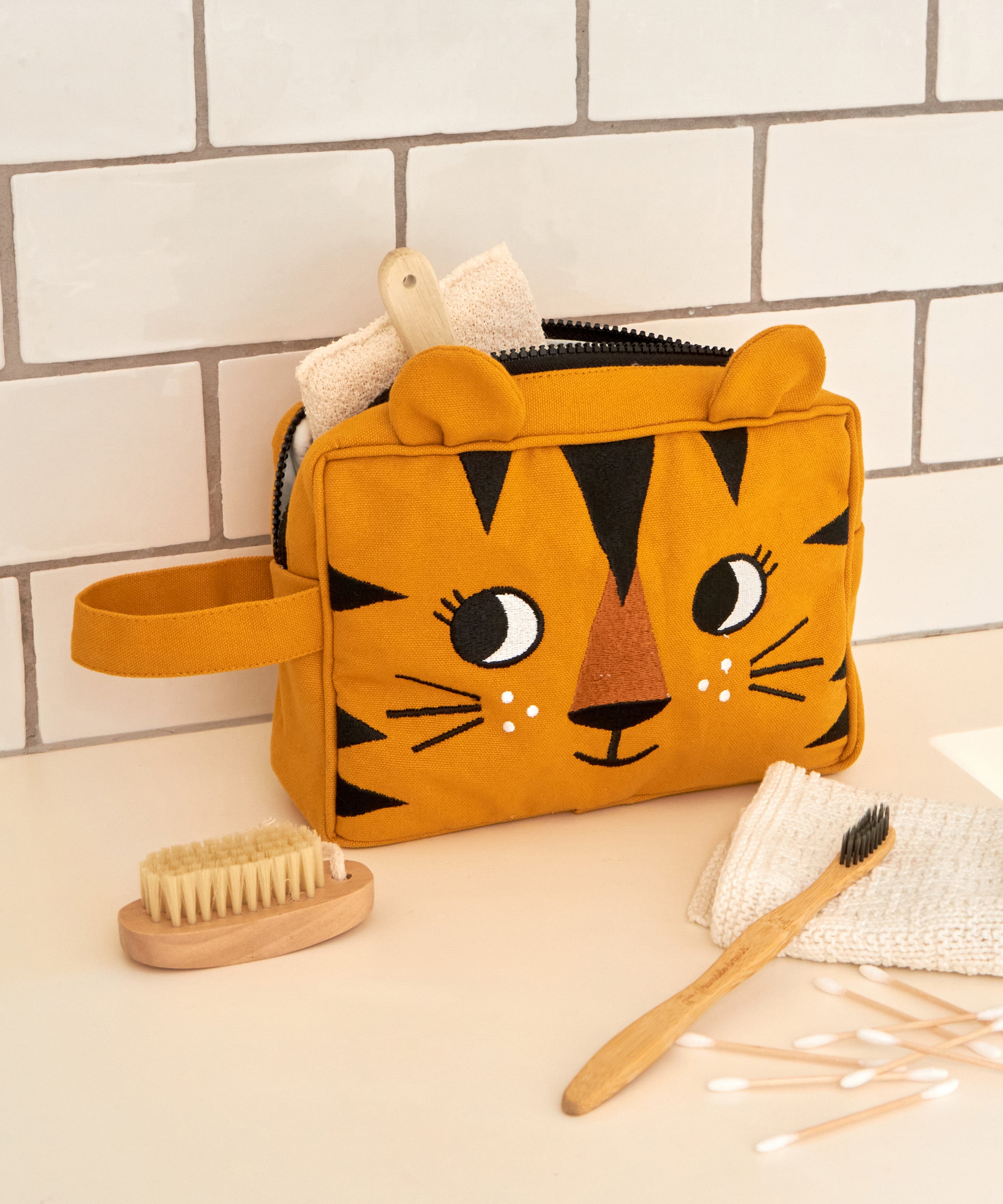 The Roommate organic cotton kids washbag, in a sunny yellow with an embroidered tiger design and 3D tiger ears. It is in a bathroom and filled with toiletries.