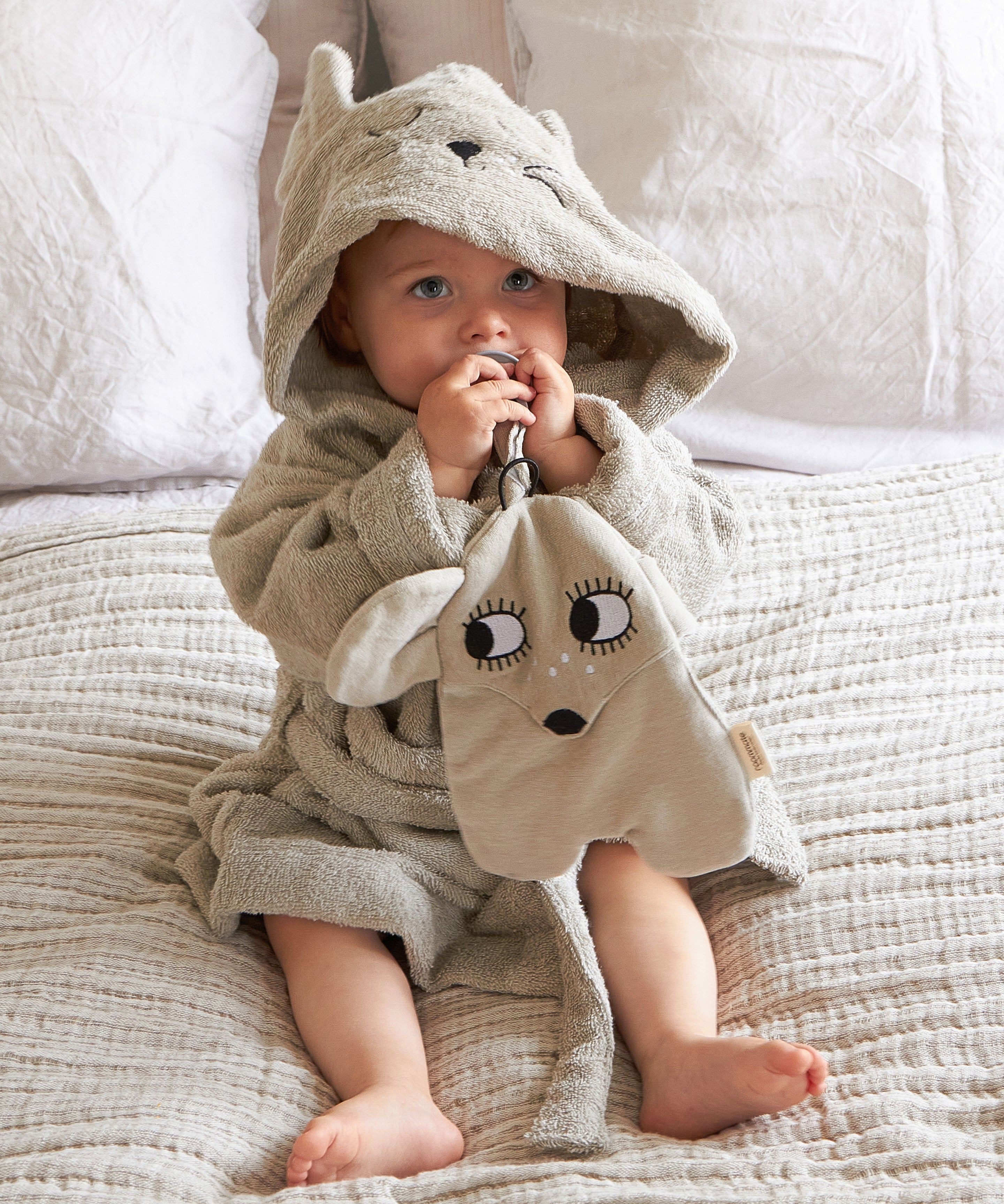 A baby wearing the Roommate grey mouse bathrobe dressing gown, and holding a Roommate comforter toy. 