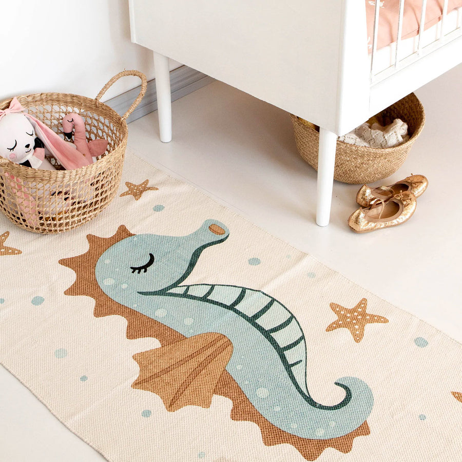 Close up of the Roommate organic cotton sea horse rug laid out on a white floor next to a basket of stuffed toys and child's bed