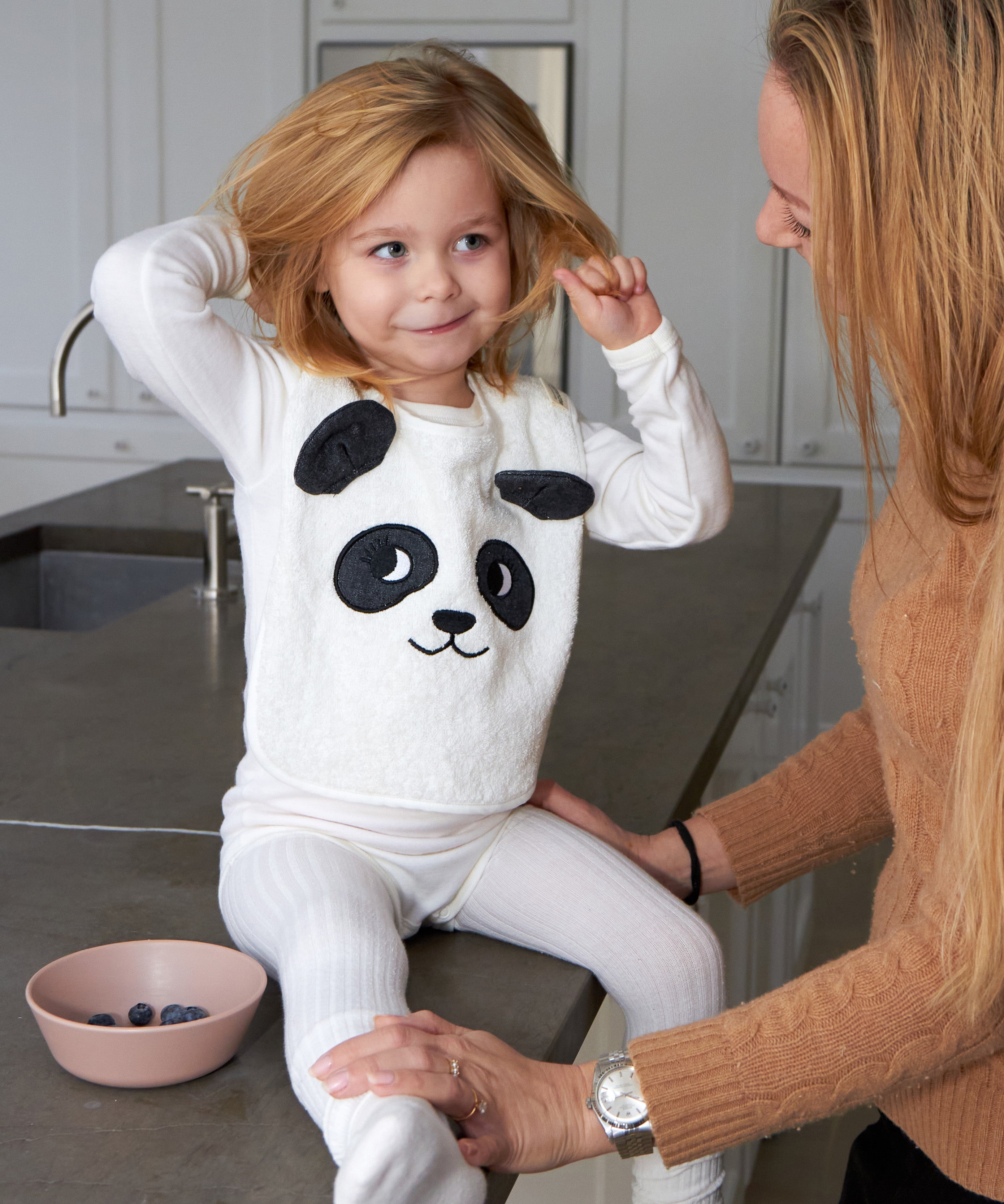 Child sat on a grey kitchen worktop wearing the Roommate Panda Baby Bib in organic cotton with embroidered panda face with 3D ears. Parent is holding her and she has a bowl of blueberries.