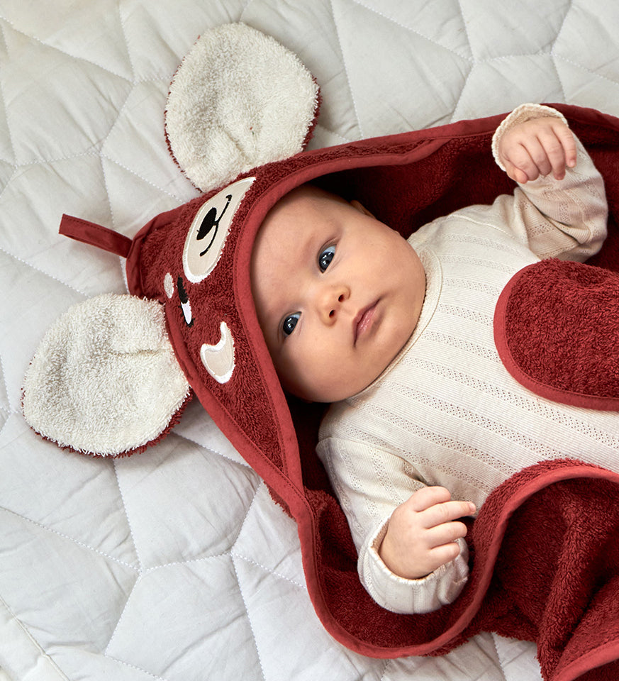 A little baby wrapped up in the Roommate organic cotton hooded towel, in red with a cute red panda face embroidered onto the hood. 