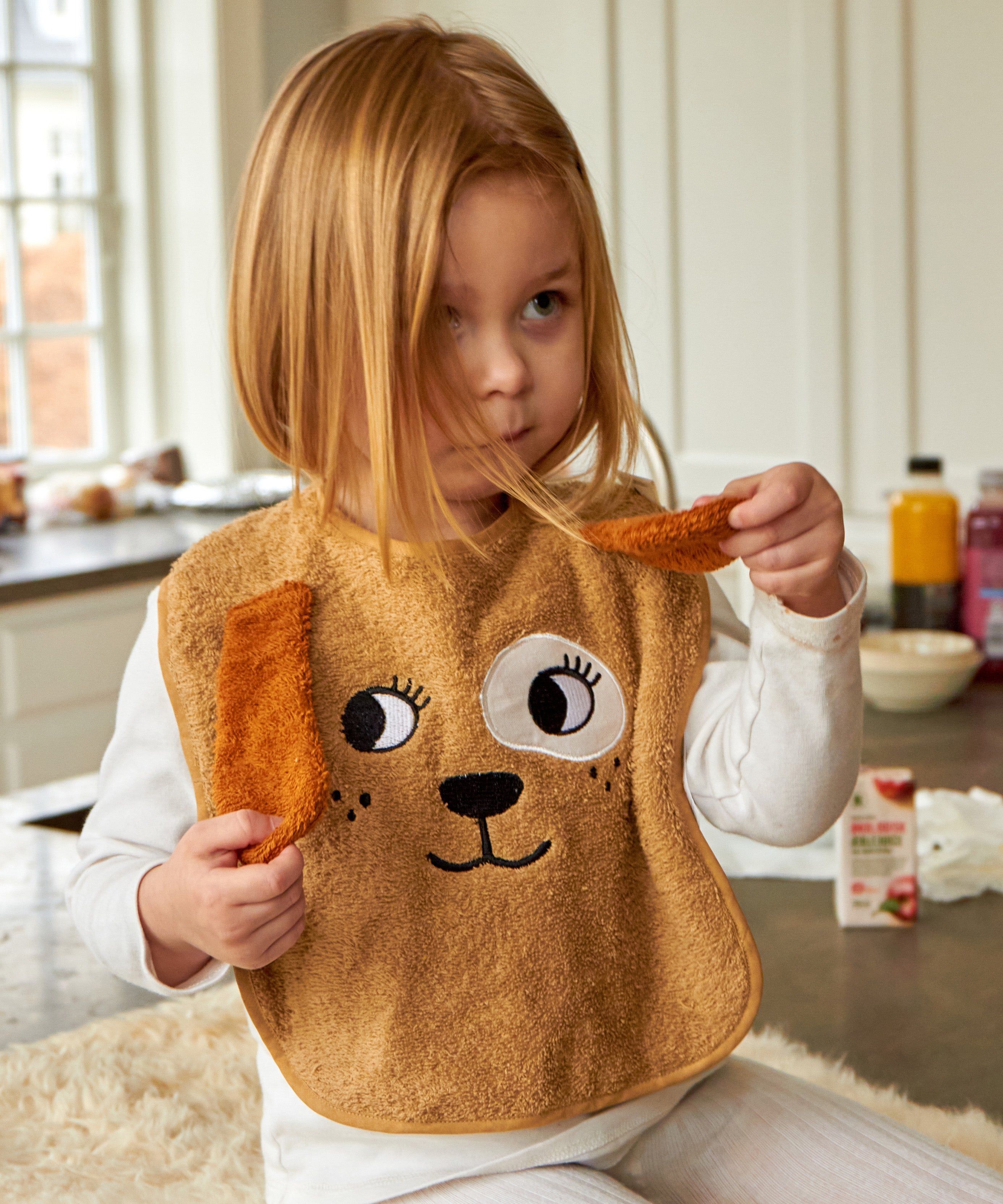 Child wearing the Roommate Dog Baby Bib with an embroidered puppy face with floppy 3D ears. She is sat on a worktop holding up the puppy ears.