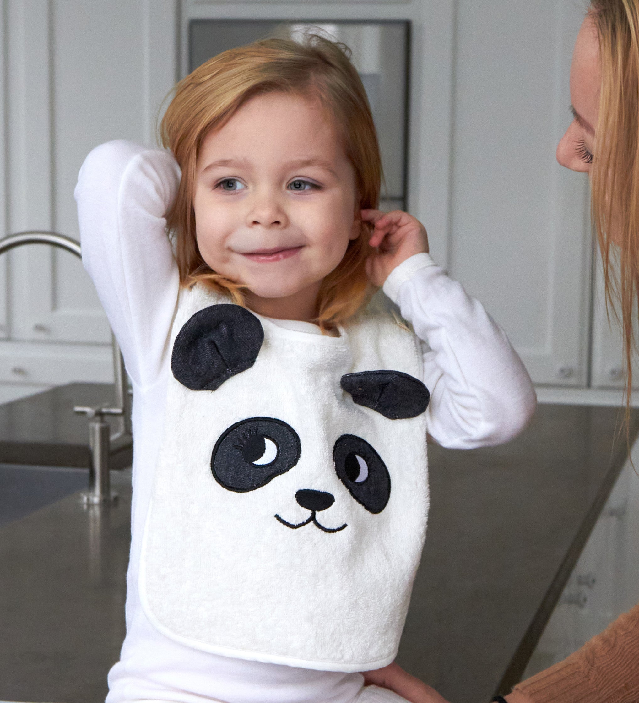 Child sat on a grey kitchen worktop wearing the Roommate Panda Baby Bib in organic cotton with embroidered panda face with 3D ears.