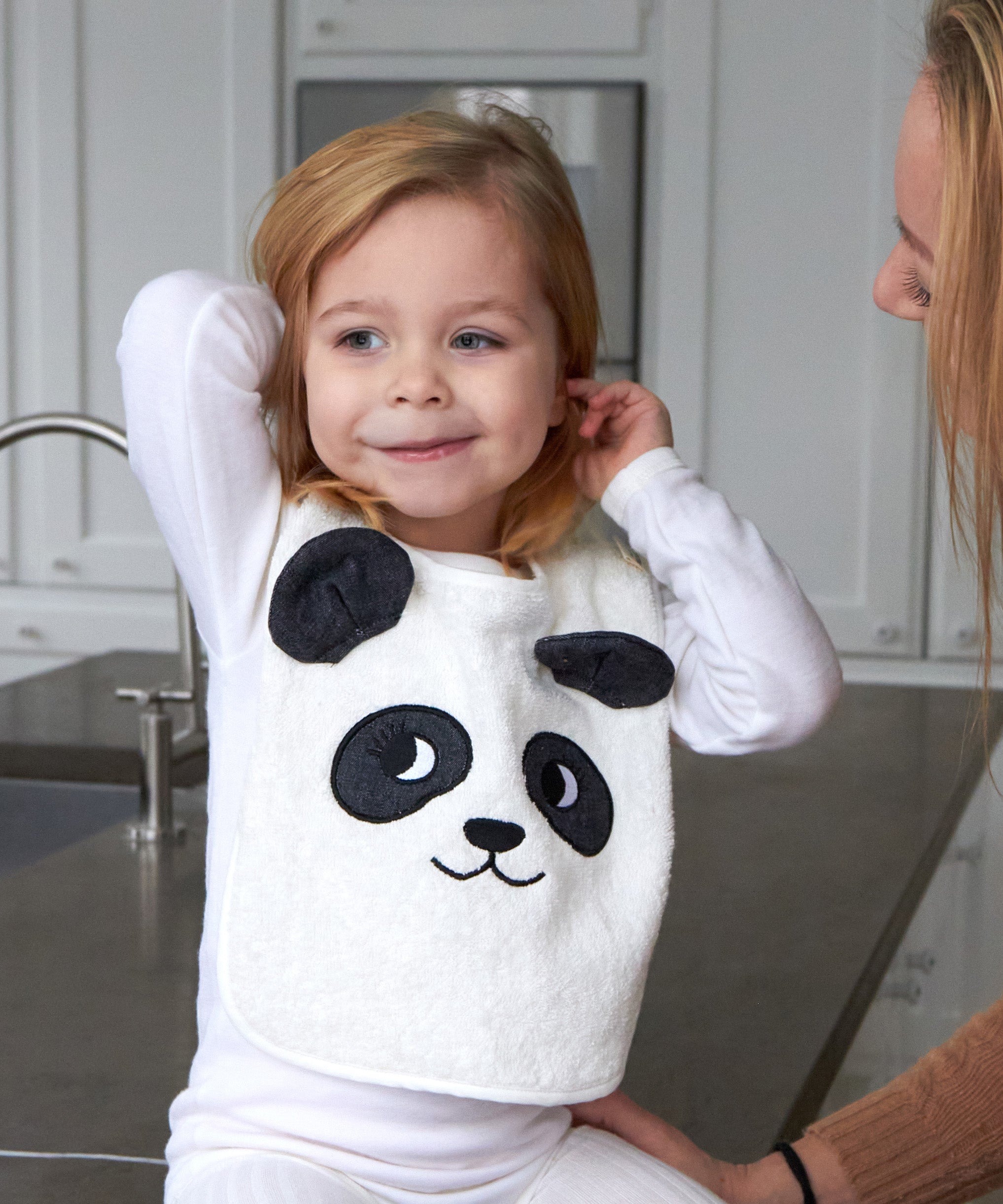 Child sat on a grey kitchen worktop wearing the Roommate Panda Baby Bib in organic cotton with embroidered panda face with 3D ears.