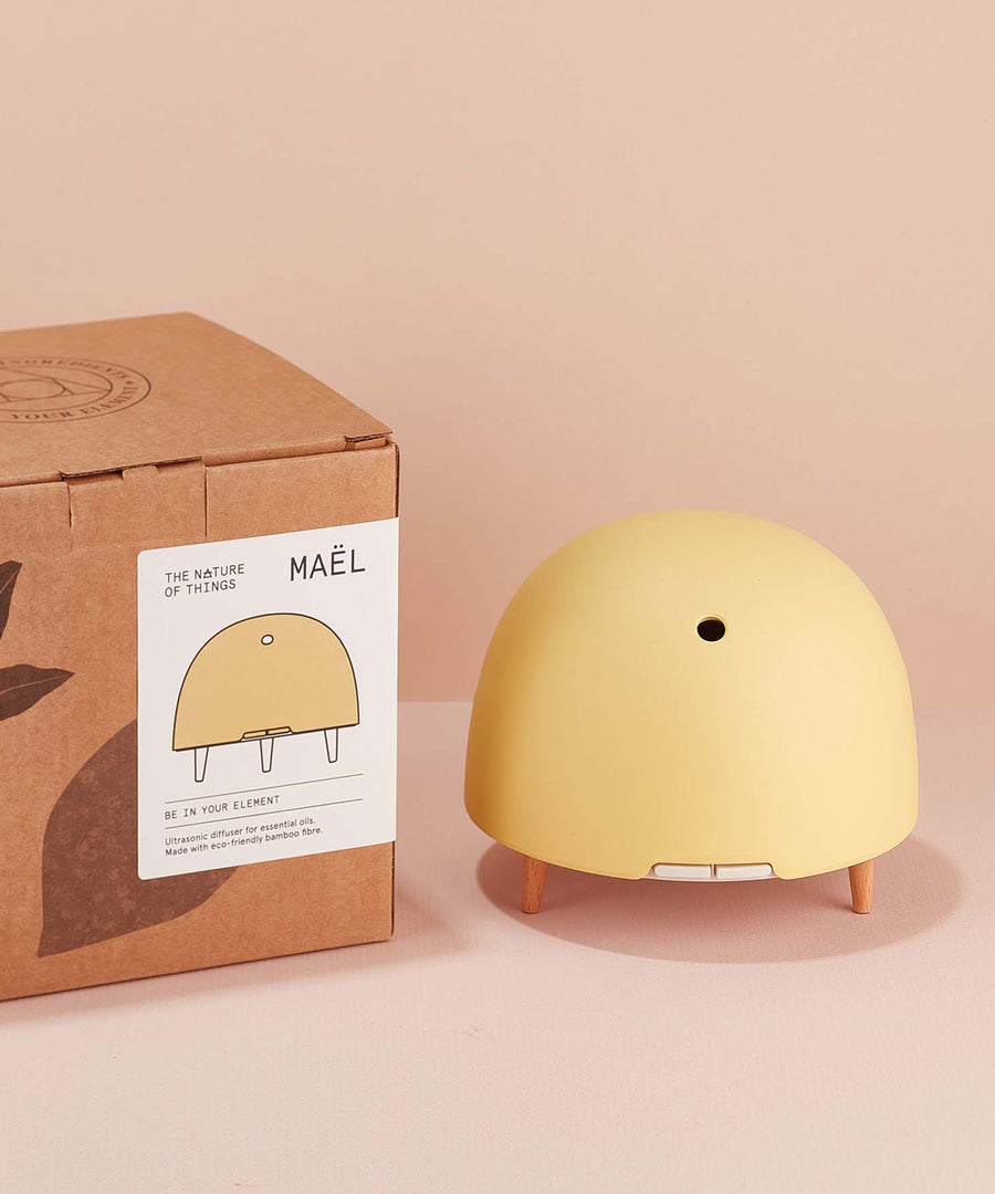 Maël Diffuser, a light yellow dome plug-in essential oil diffuser with wooden feet next to its cardboard box. 