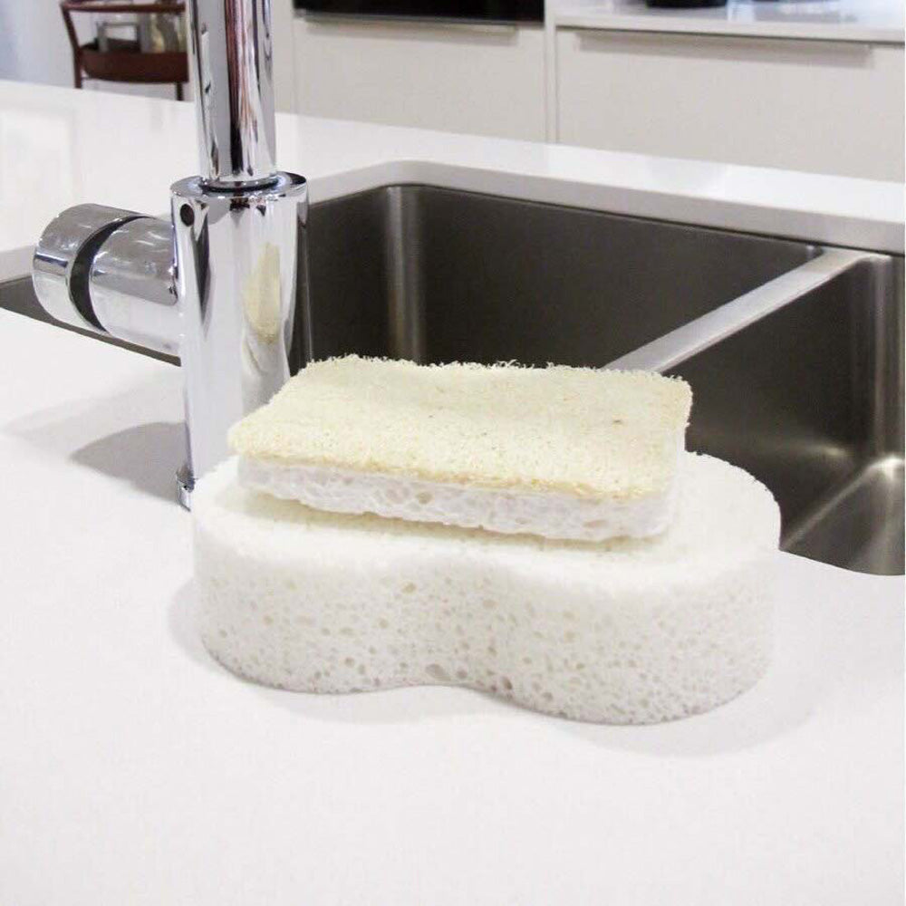 Seep natural cellulose and loofah scourer on top of a large Seep cellulose sponge next to a kitchen sink