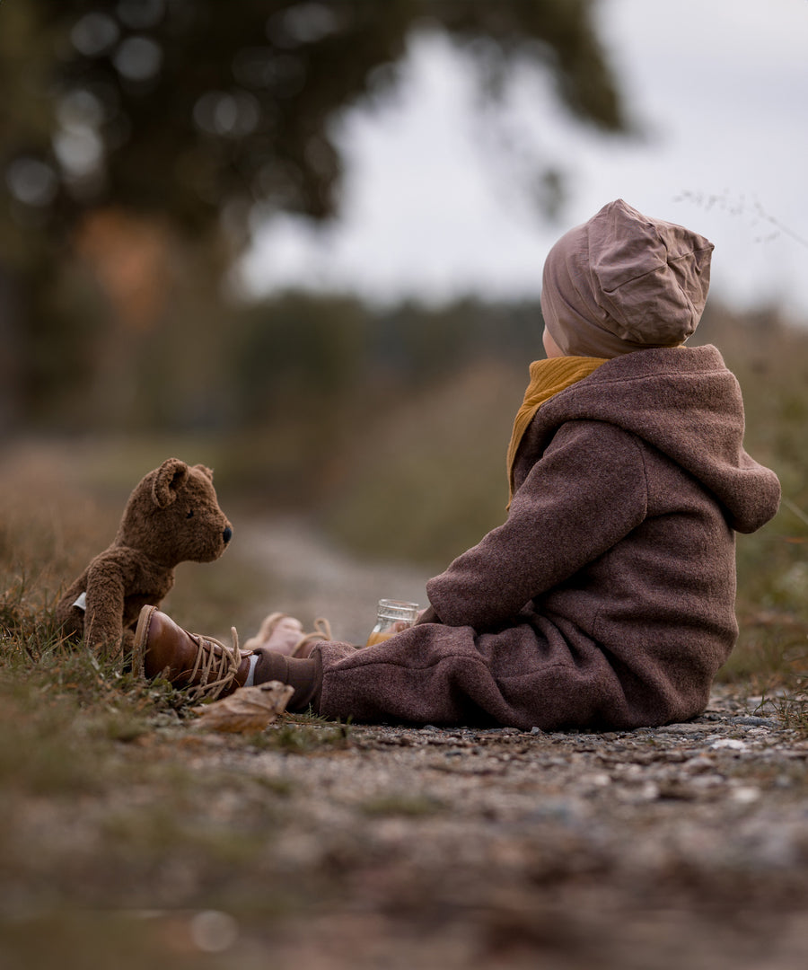 A child sitting on a gravelled surface outdoors with the Senger Large Floppy Brown Bear. 