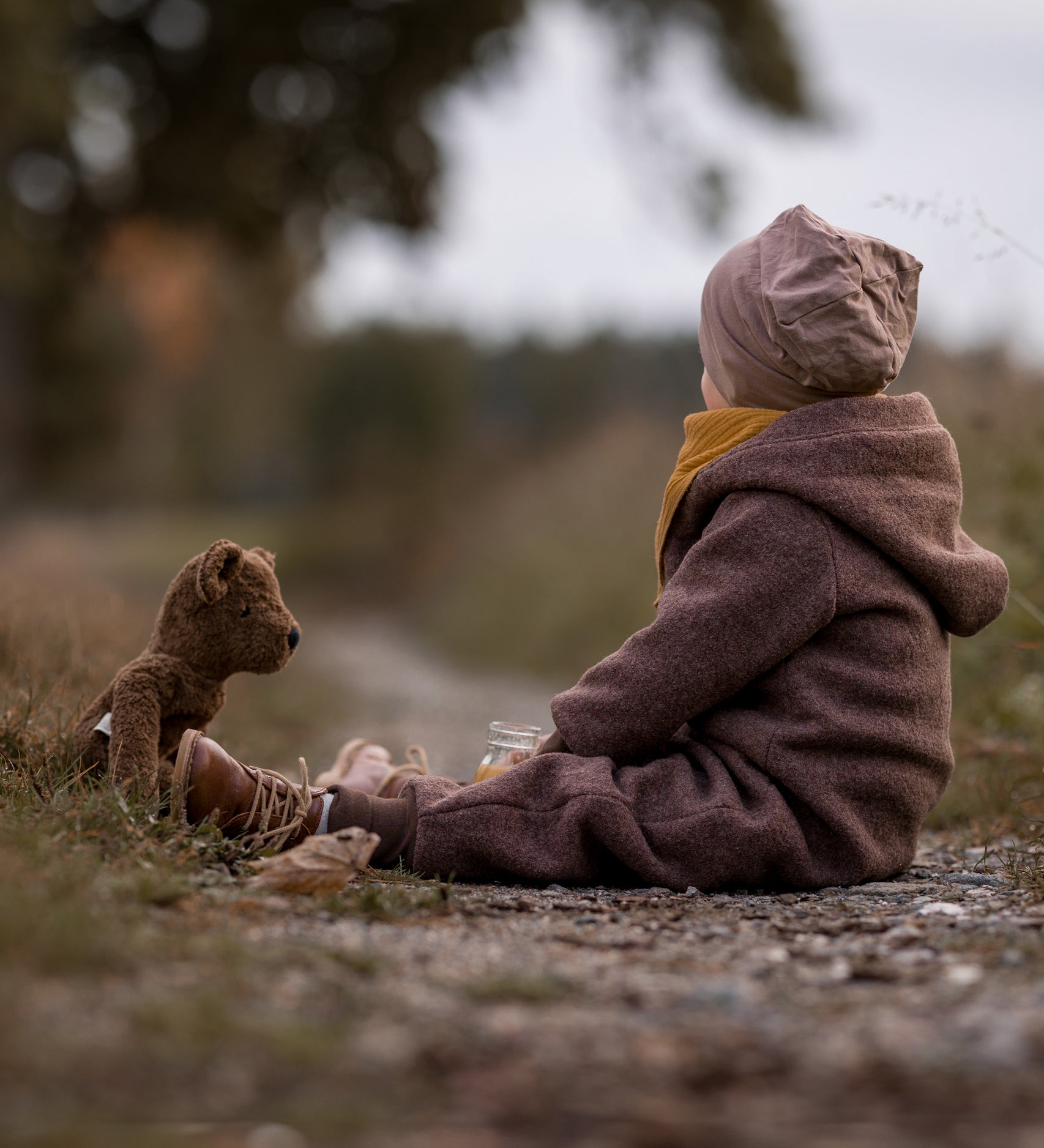 A child sitting on a gravelled surface outdoors with the Senger Large Floppy Brown Bear. 