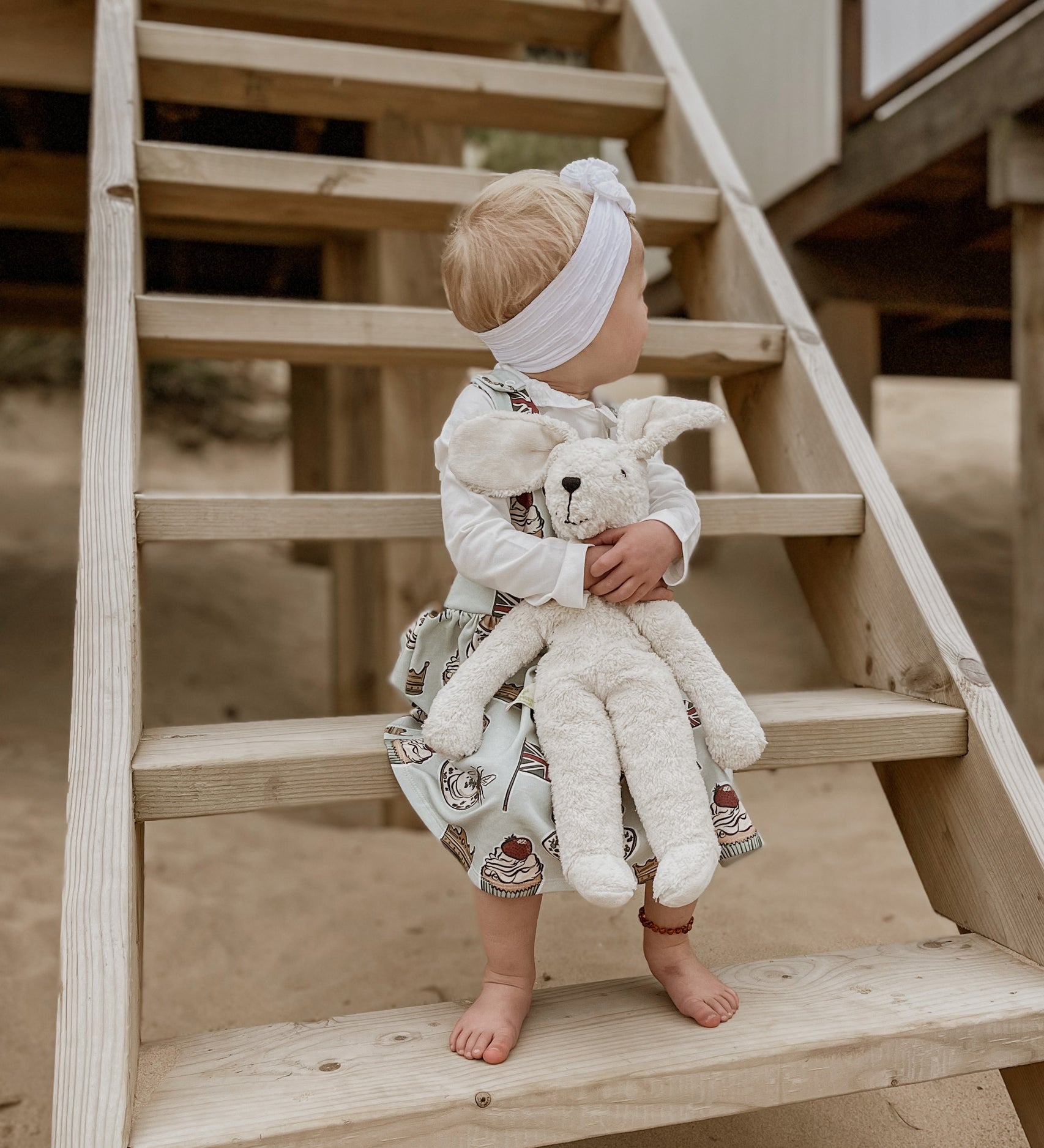 A child holding onto a Senger large floppy white rabbit with both hands. The child is sitting on wooden steps on a beach. 