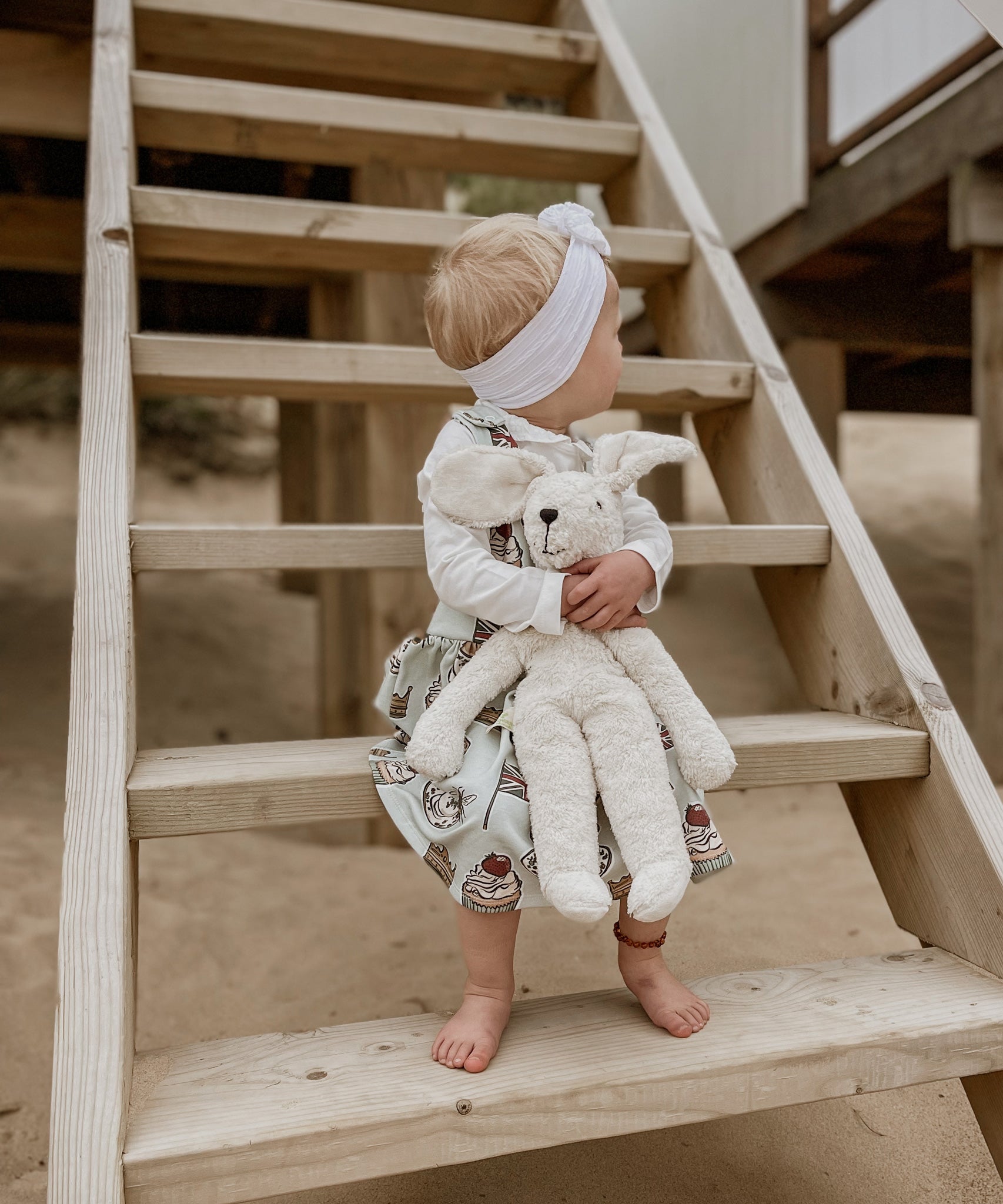 A child holding onto a Senger large floppy white rabbit with both hands. The child is sitting on wooden steps on a beach. 