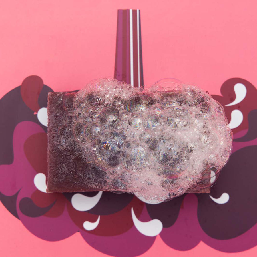 A Shower Blocks Black Cherry fragranced plastic free Gel Bar covered in a bubbly lather pictured on a pink and purple coloured patterned background