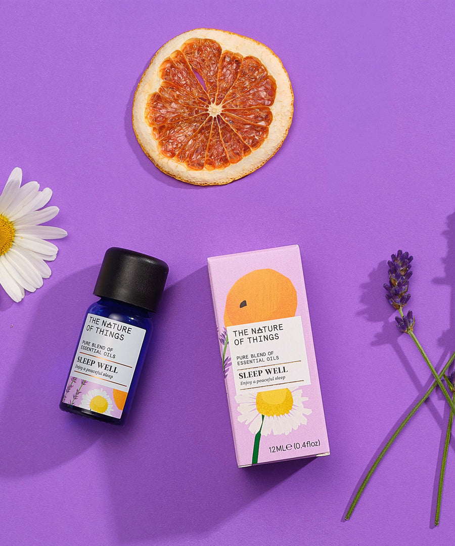 Sleep Well pure essential oil blend in a blue glass bottle next to a decorative pink cardboard box, chamomile flower, lavender flower and orange slice on a purple background. 