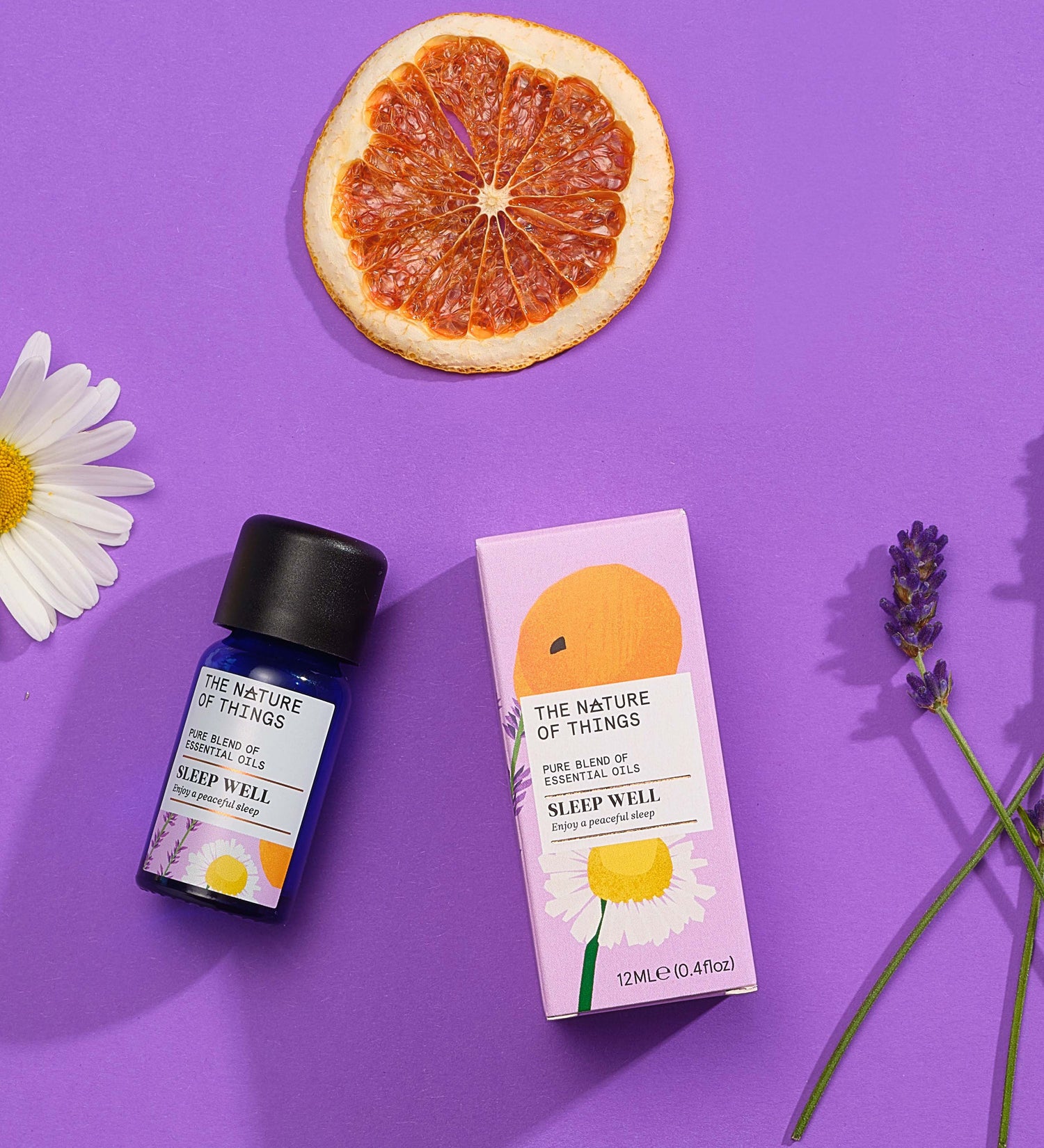 Sleep Well pure essential oil blend in a blue glass bottle next to a decorative pink cardboard box, chamomile flower, lavender flower and orange slice on a purple background. 