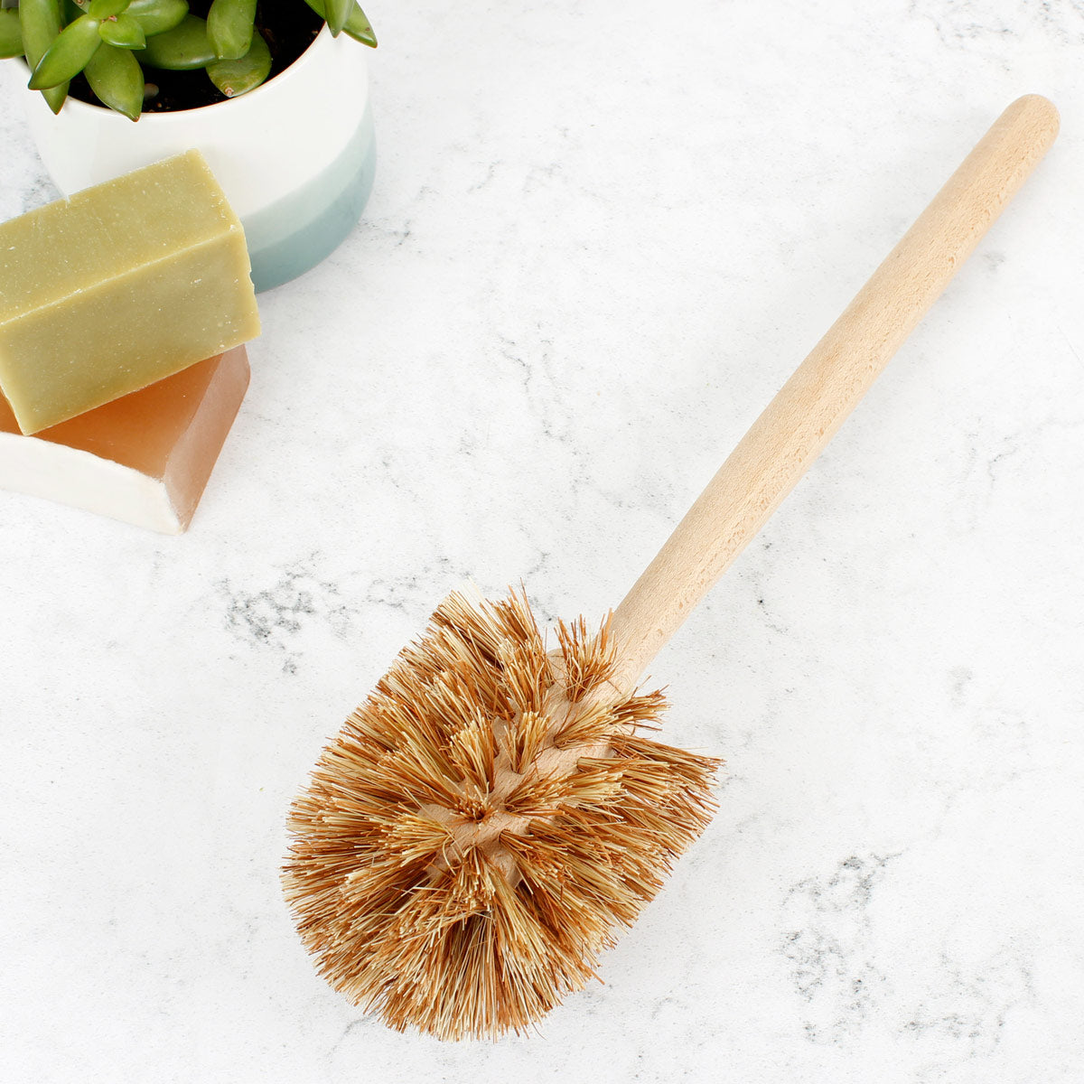 A Slice of Green plastic-free wooden toilet brush on a white background next to some bars of soap and a potted plant