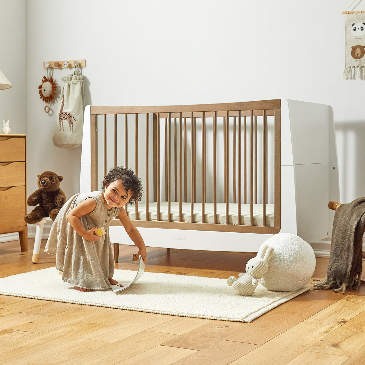 A child can be seen leaning over playing in front of a walnut SnuzKot. The child is on a white rug in a nursery