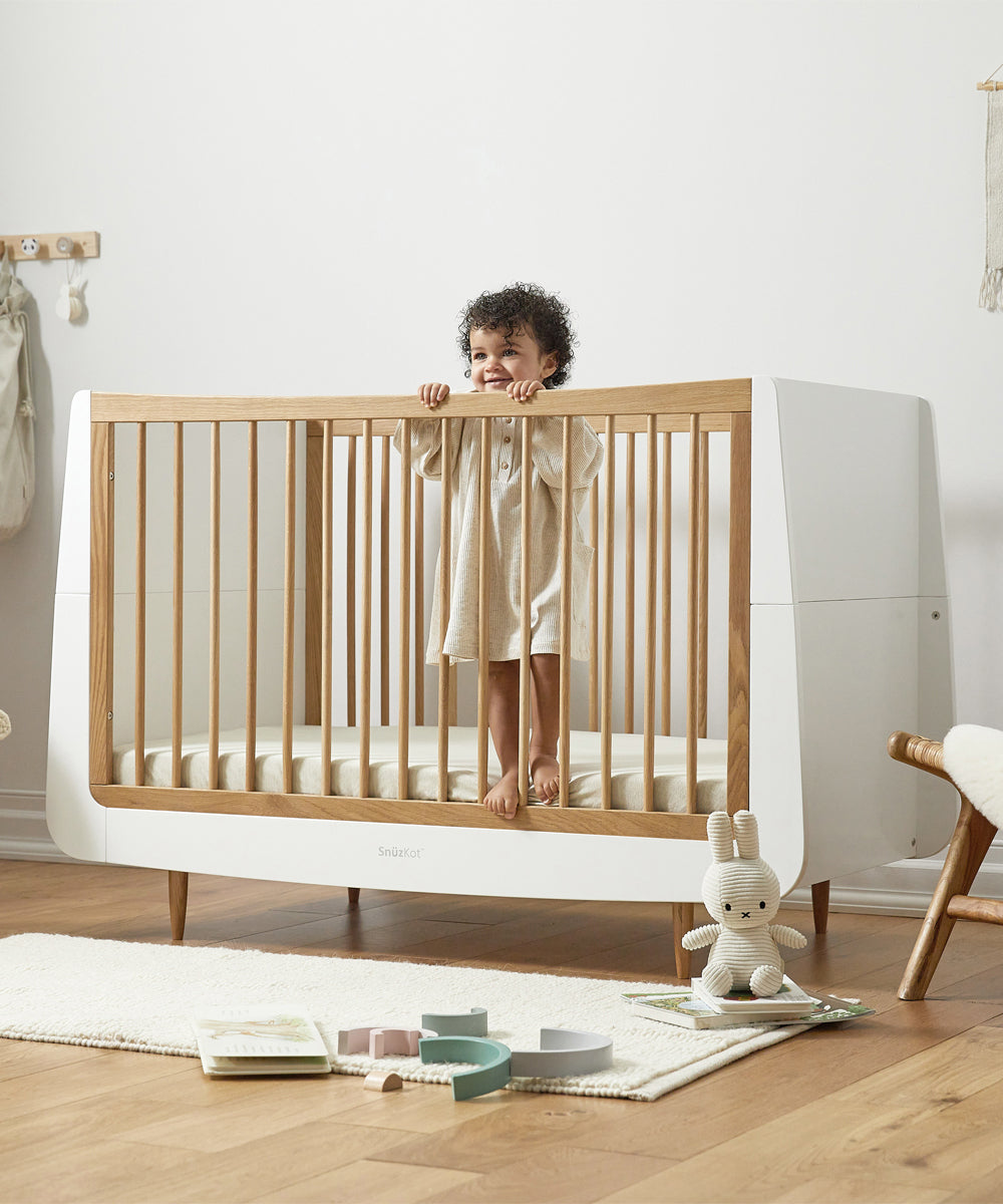 A SnuzKot Cot bed in oak pictured in a nursery. A child with curly hair can be seen standing up in the cot holding onto the side