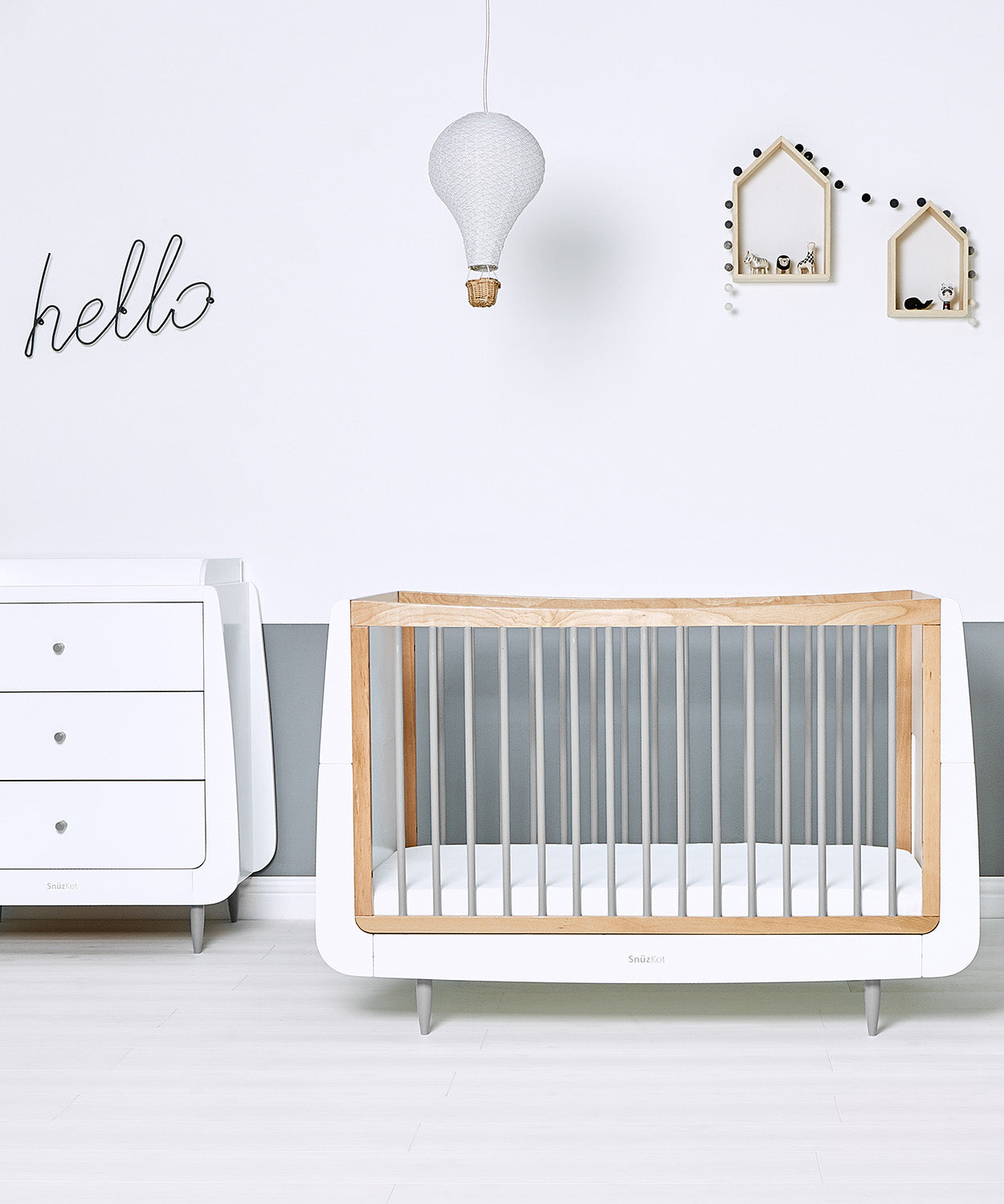 SnuzKot Skandi Cot Bed in grey with a mathcing changing table in a nursery. The nursery has white and grey coloured walls decorated with wooden house shaped shelving 