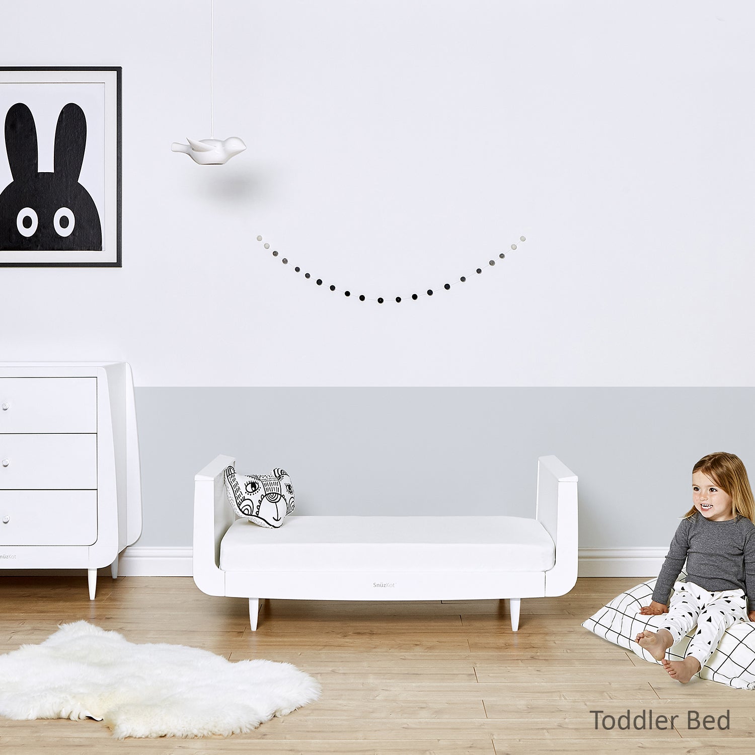 A white SnuzKot in toddler bed mode pictured in a bedroom. A child can be seen sitting on a floor cushion on the floor.