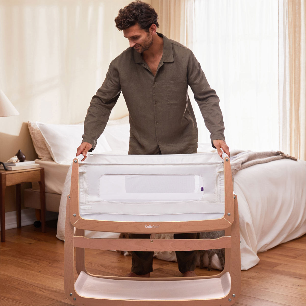 An adult can be seen lifting the bassinet of the Snuzpod 4 off the frame. There is a bed and bedside table in the background 