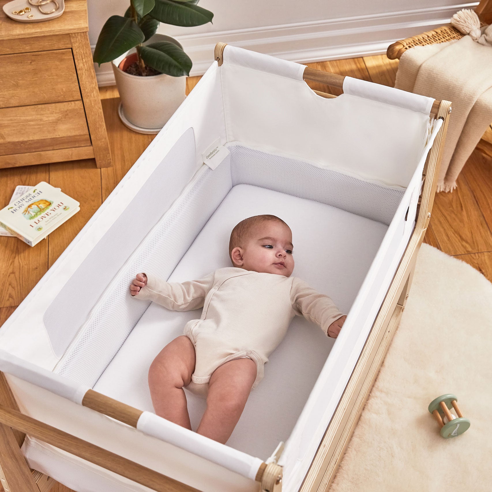 A baby is seen lying down in an oak SnuzPod 4. The Snuzpod has been placed on a light coloured rug. Some books and toys can be seen on the floor 