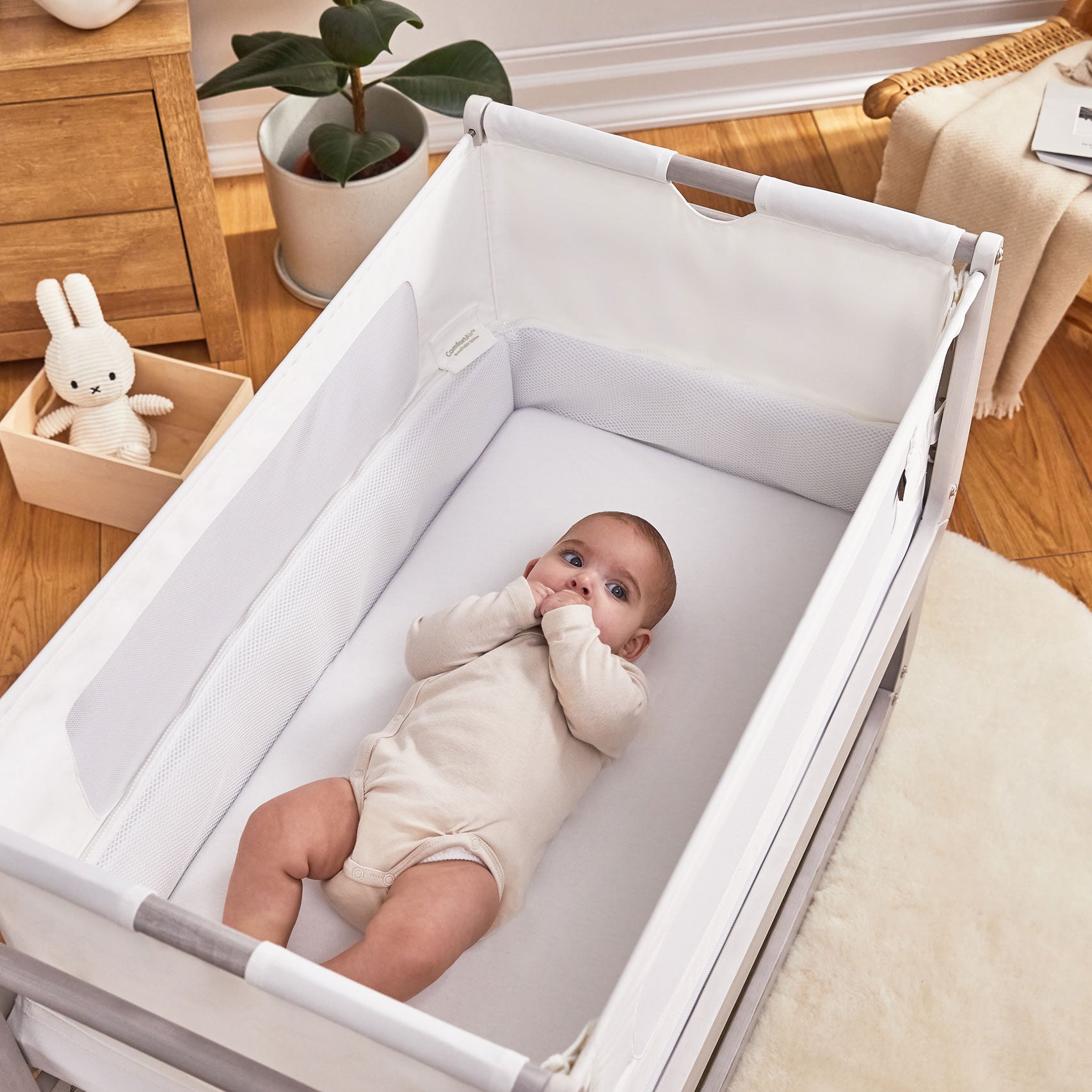 A baby is lying down in a silver birch coloured SnuzPod 4. The crib is placed on a light coloured rug, a soft toy can be seen on the floor besides the crib 