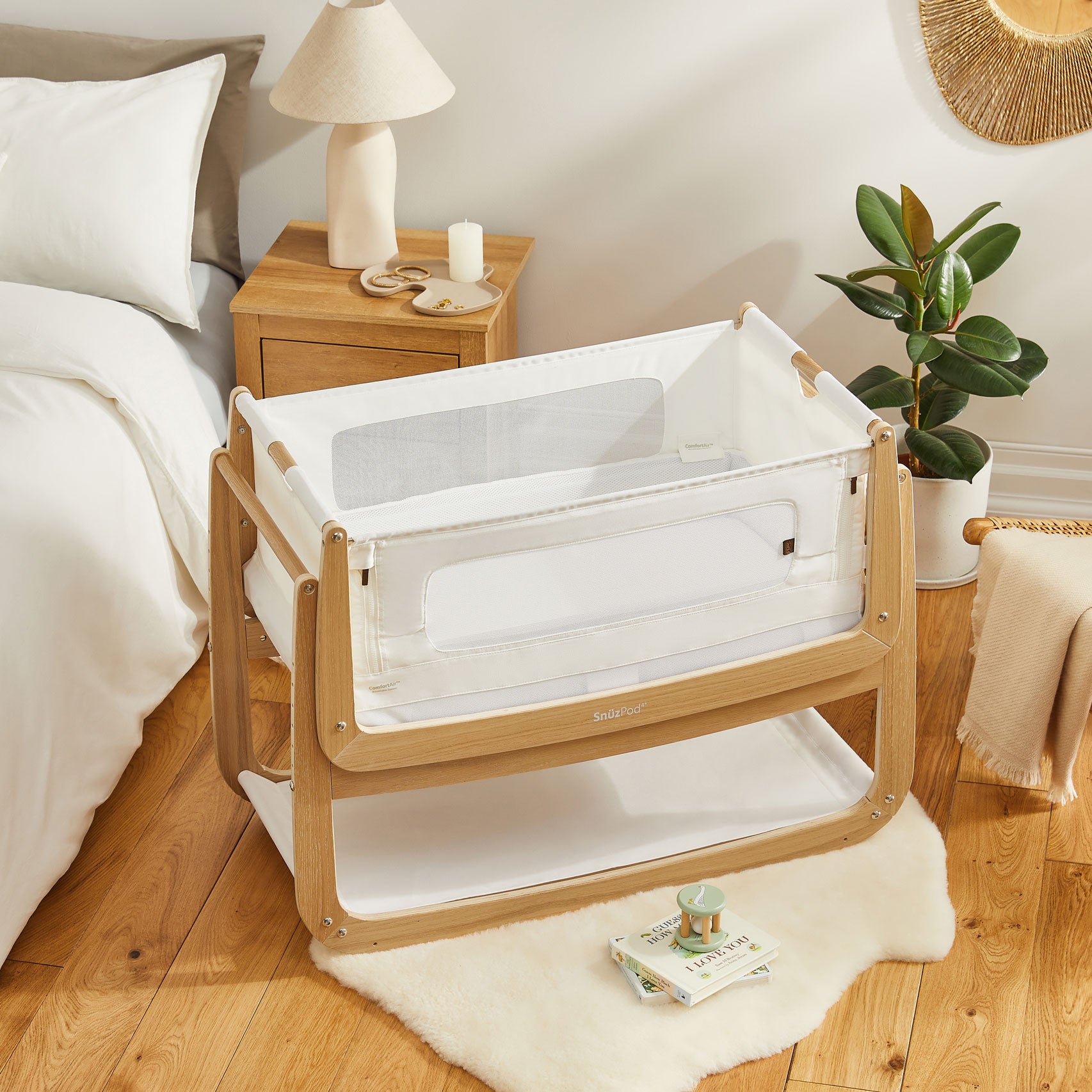 An oak SnuzPod bedside crib placed on a light coloured rug. A bed, bedside table and a house plant can be seen in the background