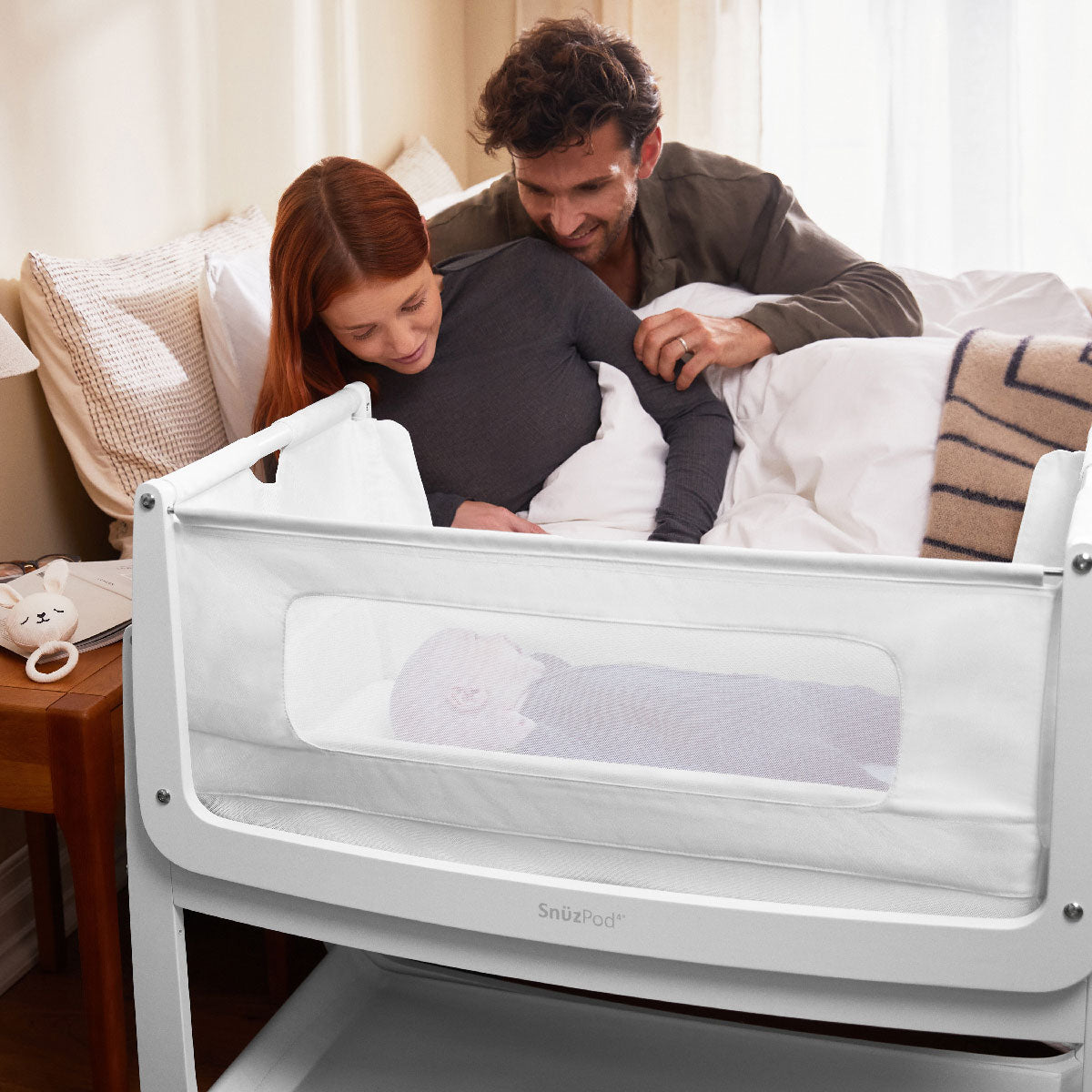 A white coloured SnuzPod placed next to a bed with a baby lying inside. There are two adults on the bed behind looking down at the baby 