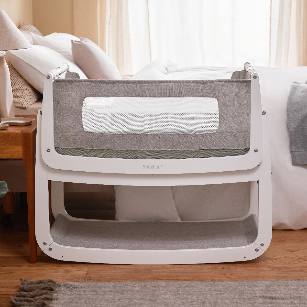 A Snuzpod 4 placed next to bad, the Snuzpod has a white colured frame with a grey coloured liner and material 