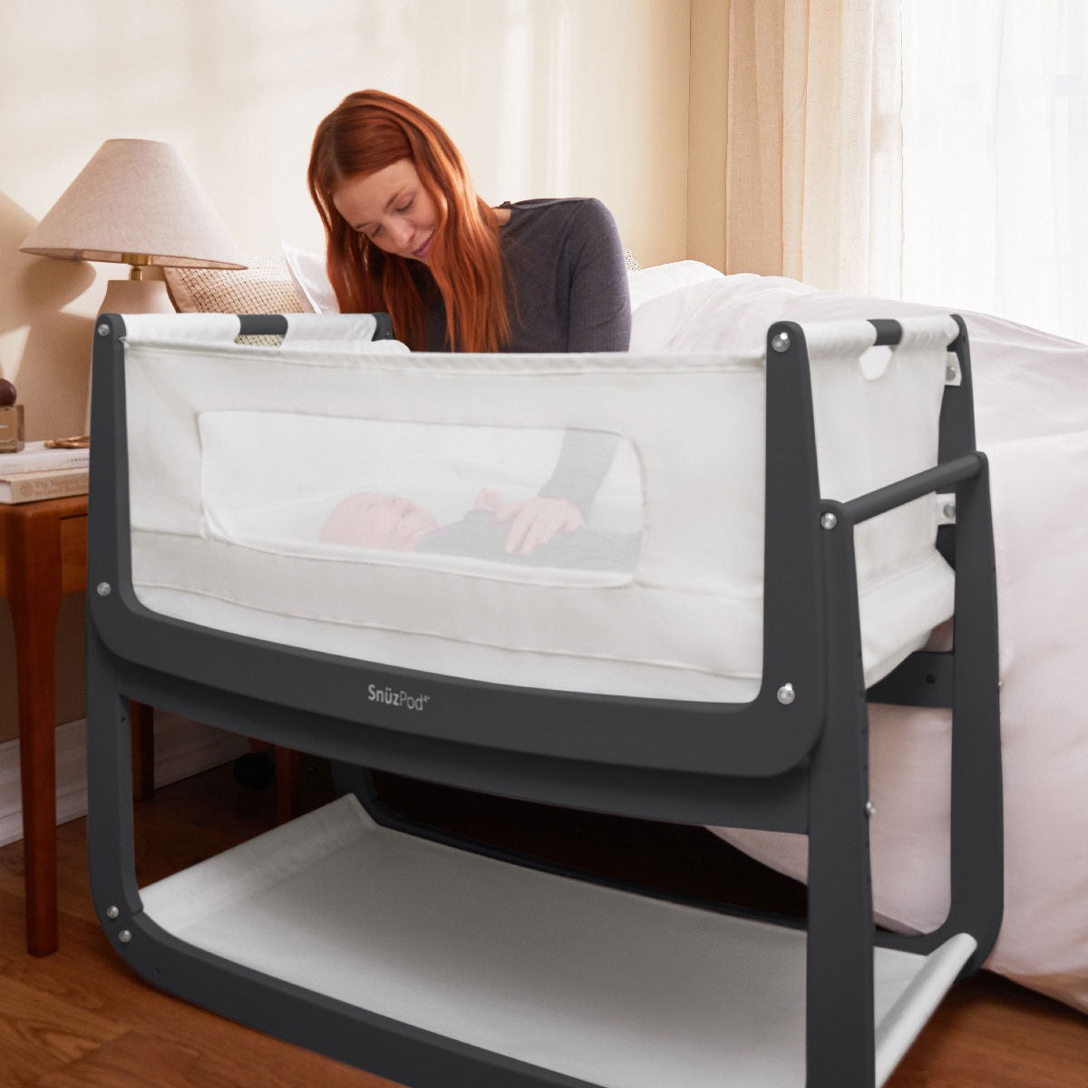 A slate grey coloured SnuzPod 4 placed next to a bed. A baby can be seen lying inside with an adult looking over them 