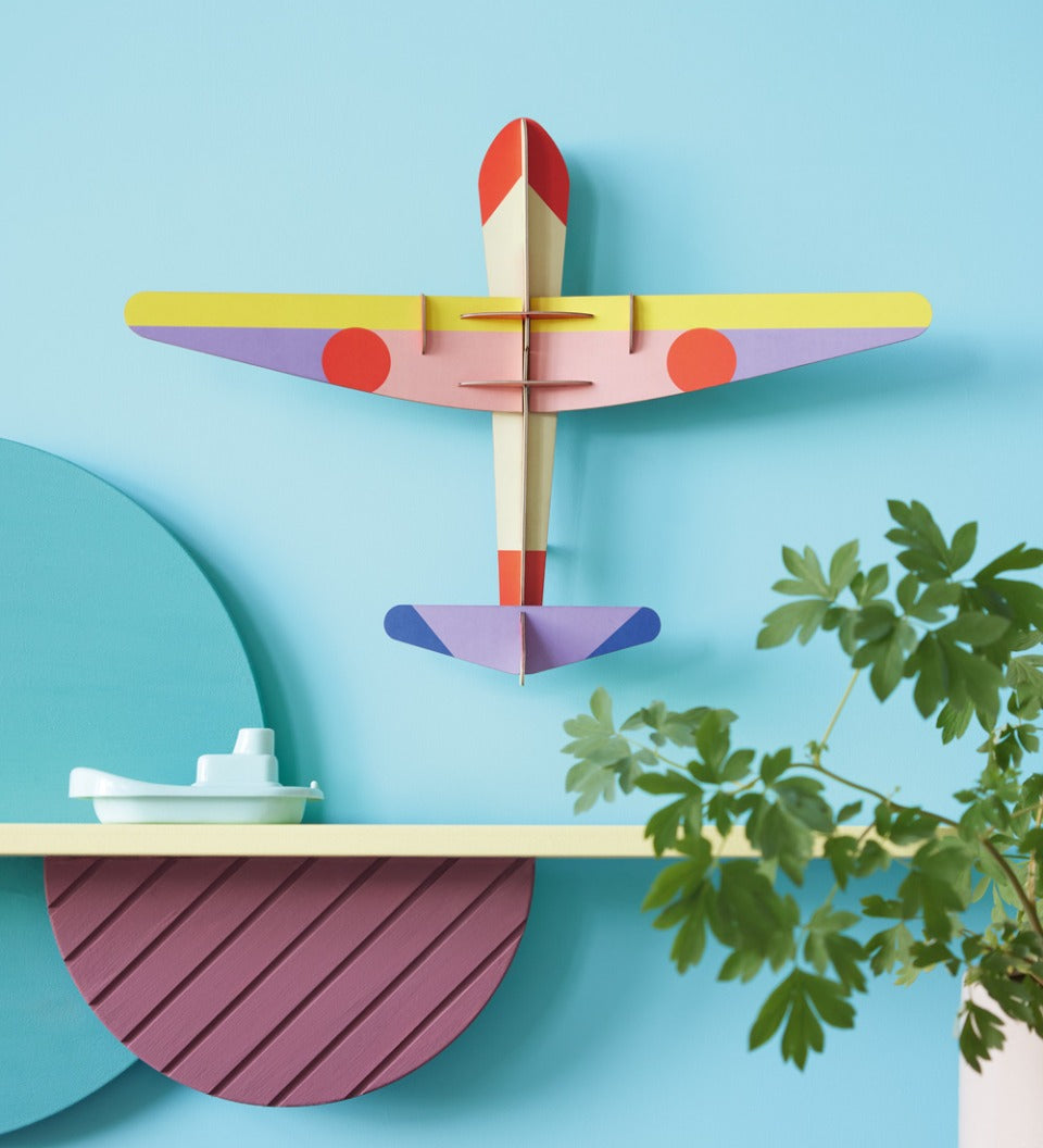 Studio Roof DIY card Plane wall decoration on a blue wall above a yellow shelf and a potted plant