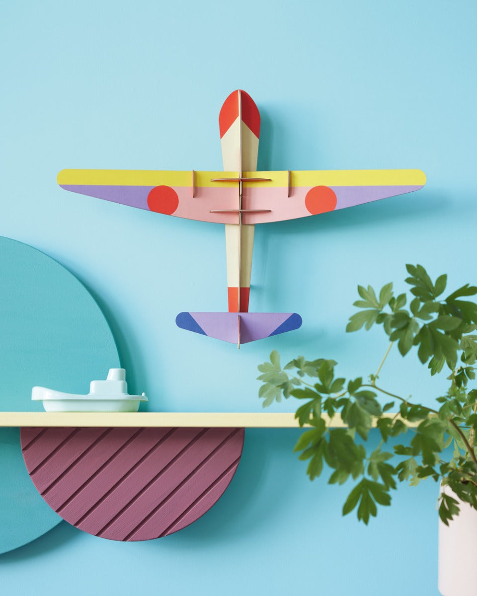 Studio Roof DIY card Plane wall decoration on a blue wall above a yellow shelf and a potted plant