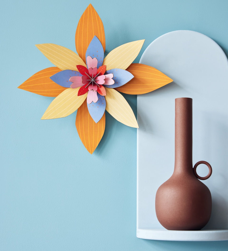 Studio Roof orange and blue craft flower hanging on a blue wall next to a light blue shelf with a brown candle holder