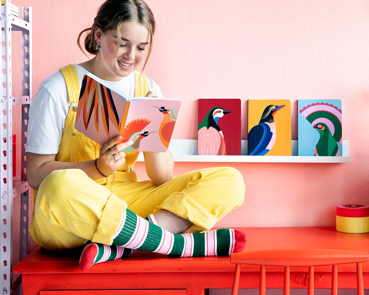 Girl sat on top of a red desk writing inside a Studio Roof Paradise Bird eco-friendly Notebook