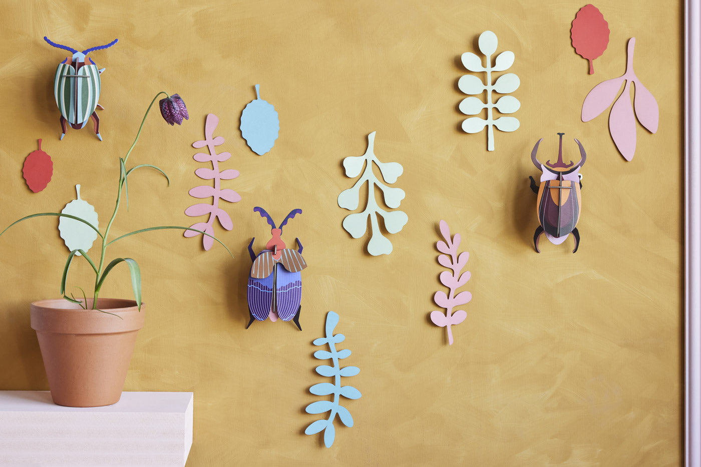 Studio Roof card craft bugs hanging on a yellow wall next to a potted plant