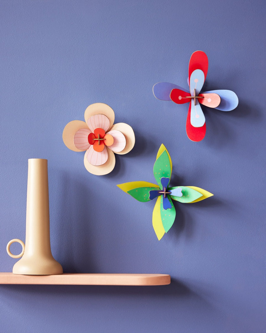 3 Studio Roof craft flowers hung on a purple wall next to a beige shelf and candle holder